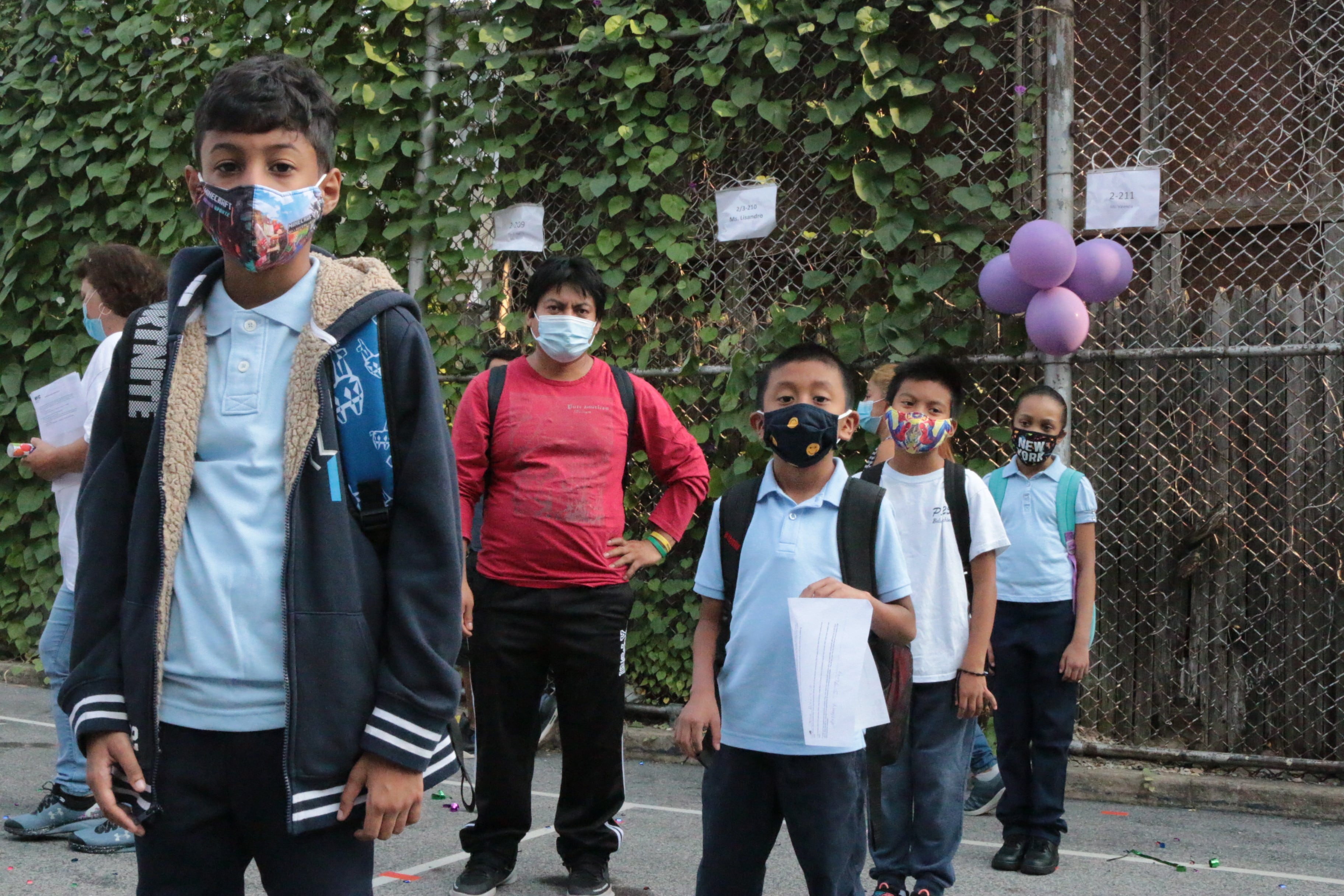 Students stand in a line next to a fence on the first day of school, each wearing protective masks.
