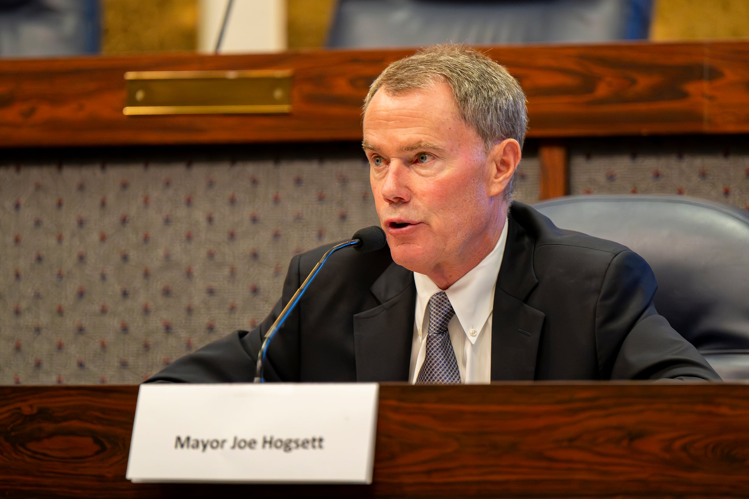 A photograph of a white man in a suit sitting at a wooden desk speaking into a microphone.