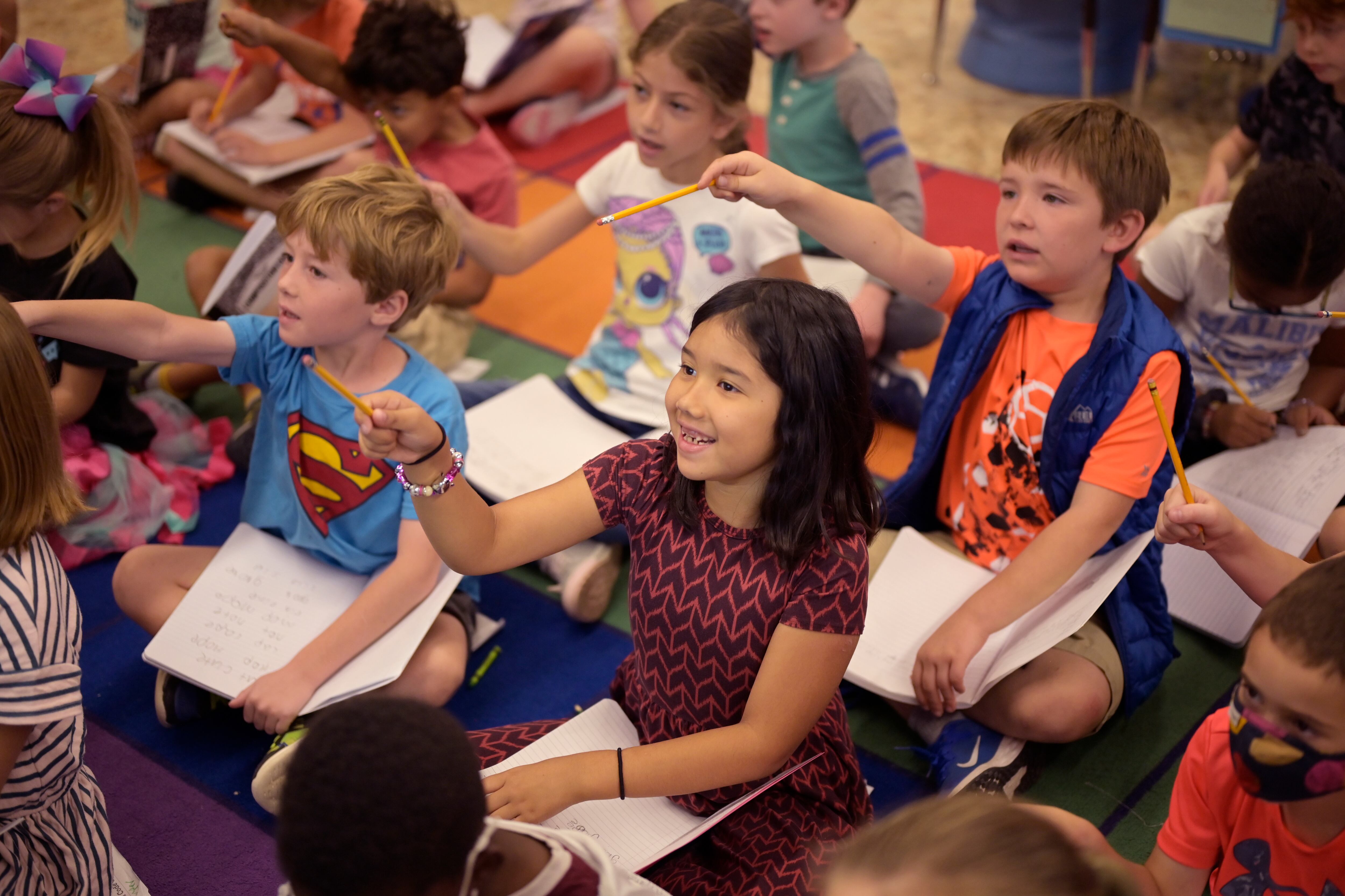A girl in a pink and black dress points her pencil towards the front of the classroom during a reading lesson.