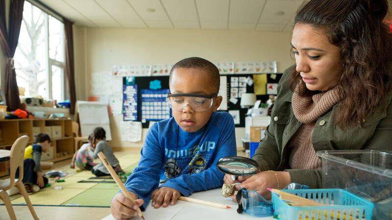 A teacher sits at a table with a kindergarten boy who is looking at a paper using a magnifying glass.