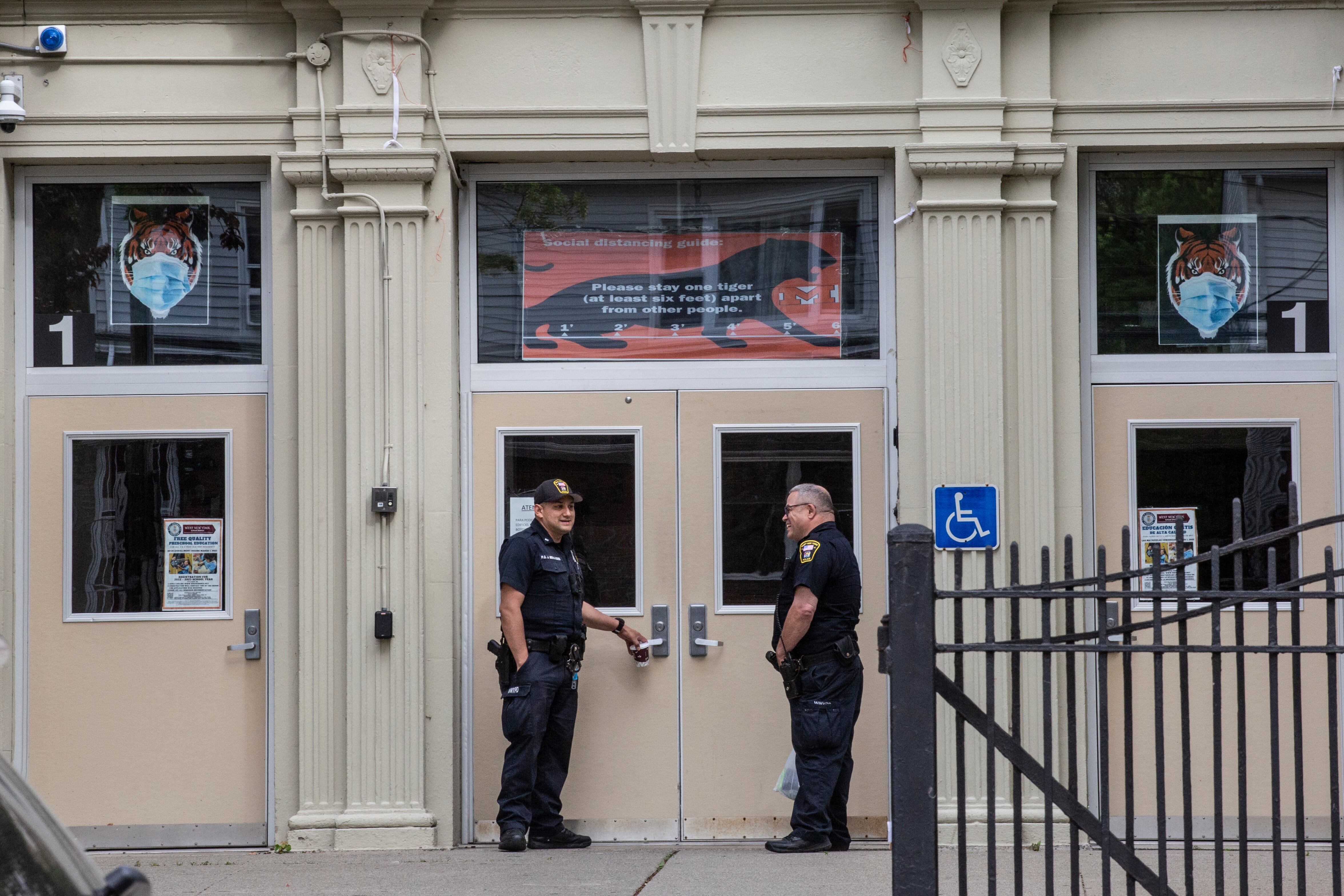 Two police officers stand in front of the entrance of a high school, the doors seen behind a metal fence.