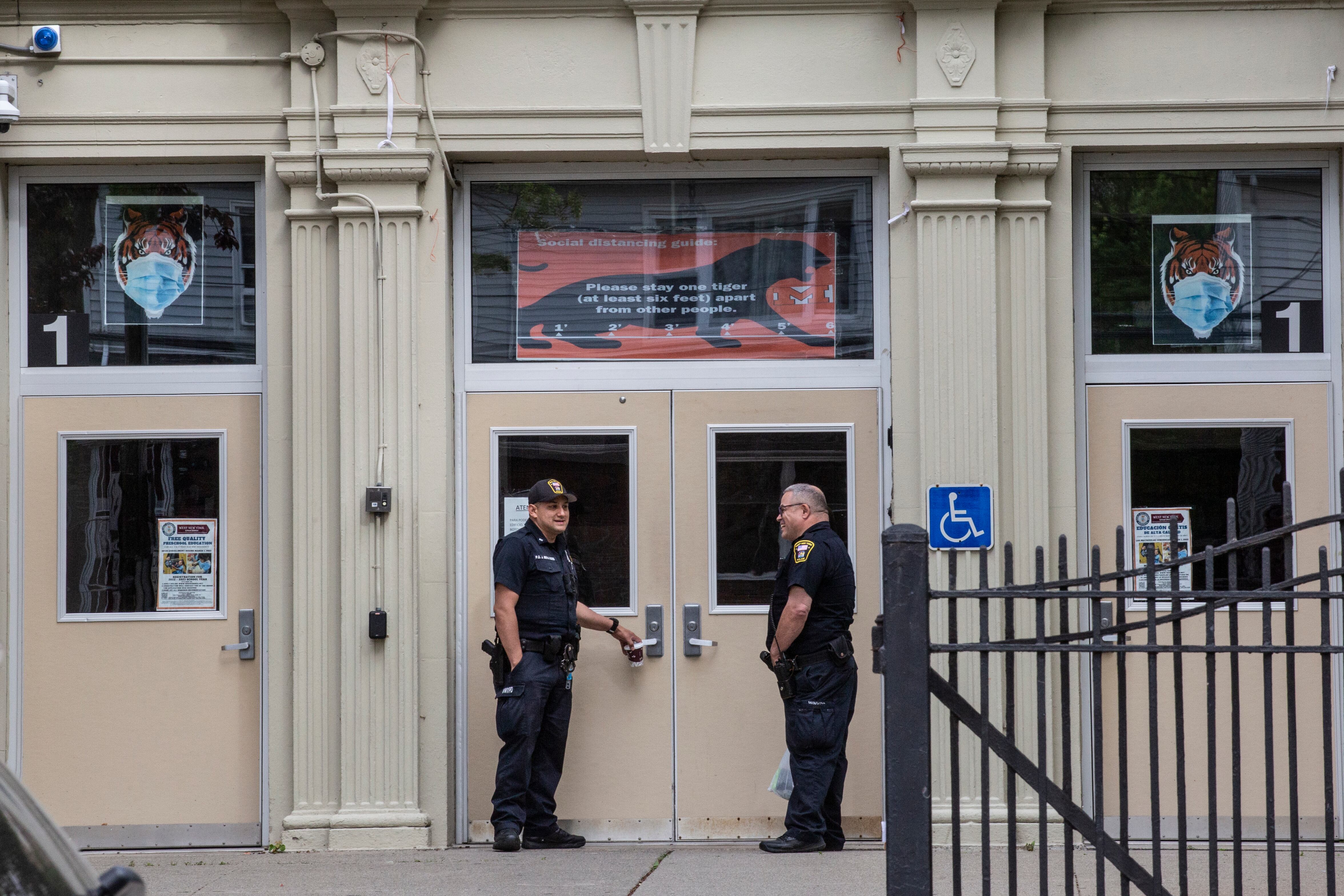 Two police officers stand in front of the entrance of a high school, the doors seen behind a metal fence.