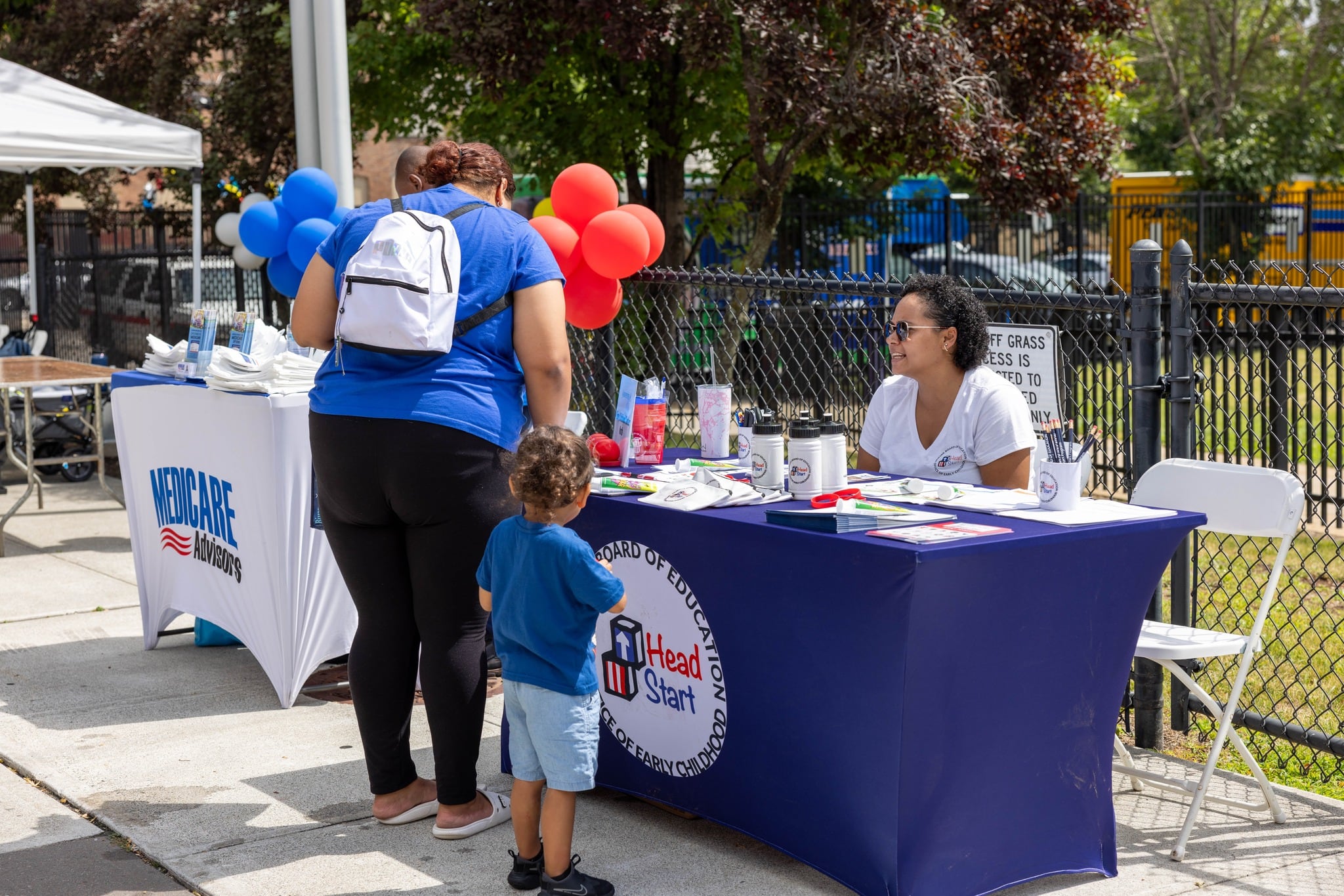 A woman wearing a backpack and a blue shirt stands next to her son as she talks with a woman sitting at a desk.