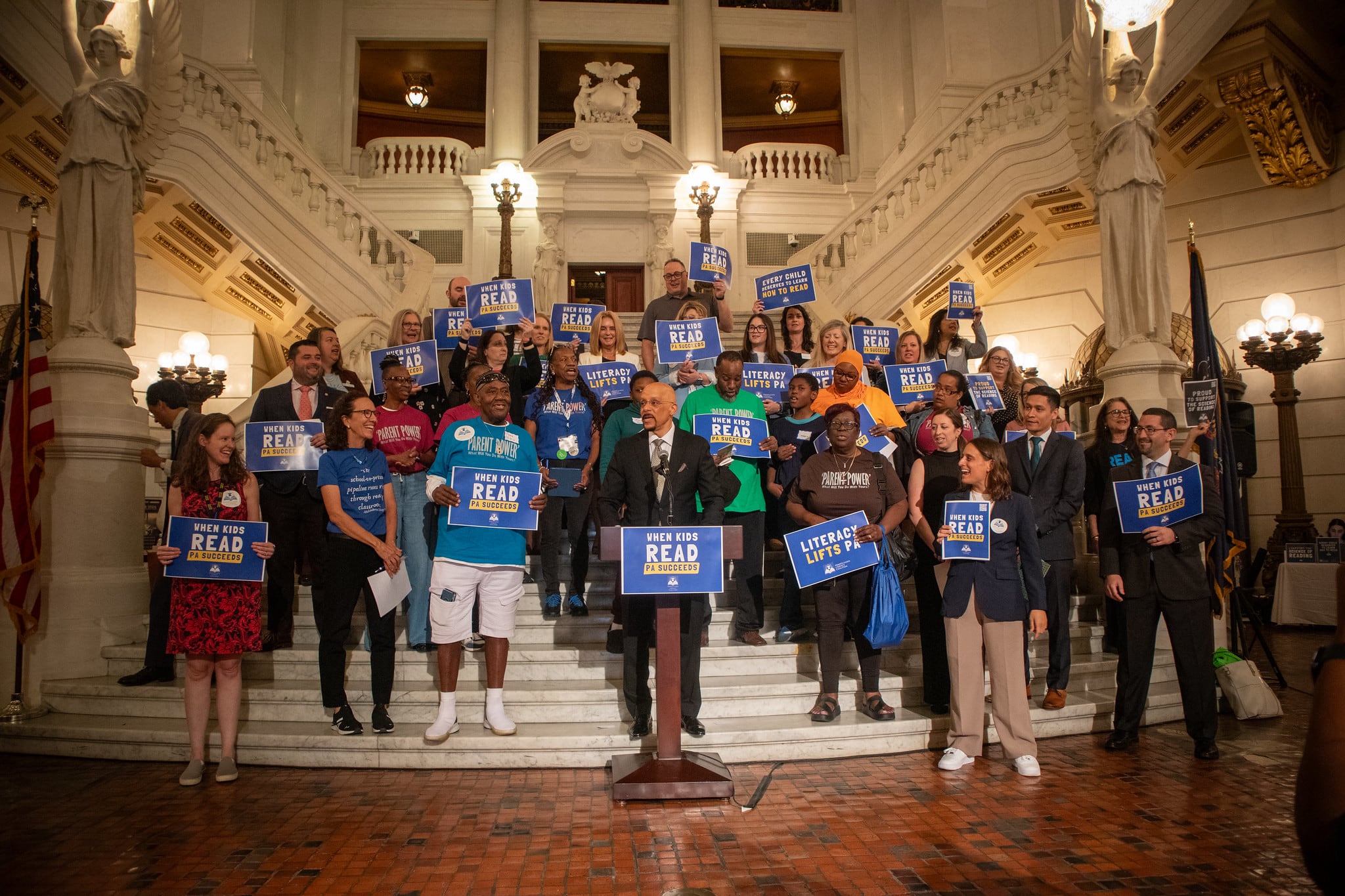 A man speaks at a podium surrounded by people with signs.