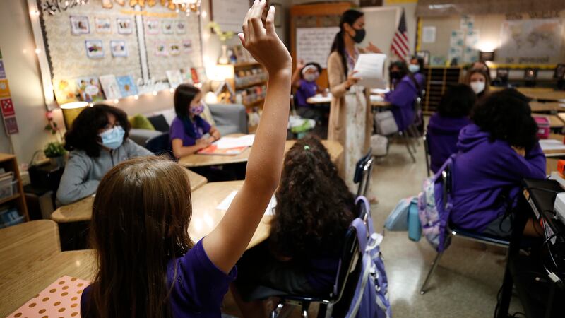A student raises their hand in a classroom, students all wearing purple and sitting at circular group desks.
