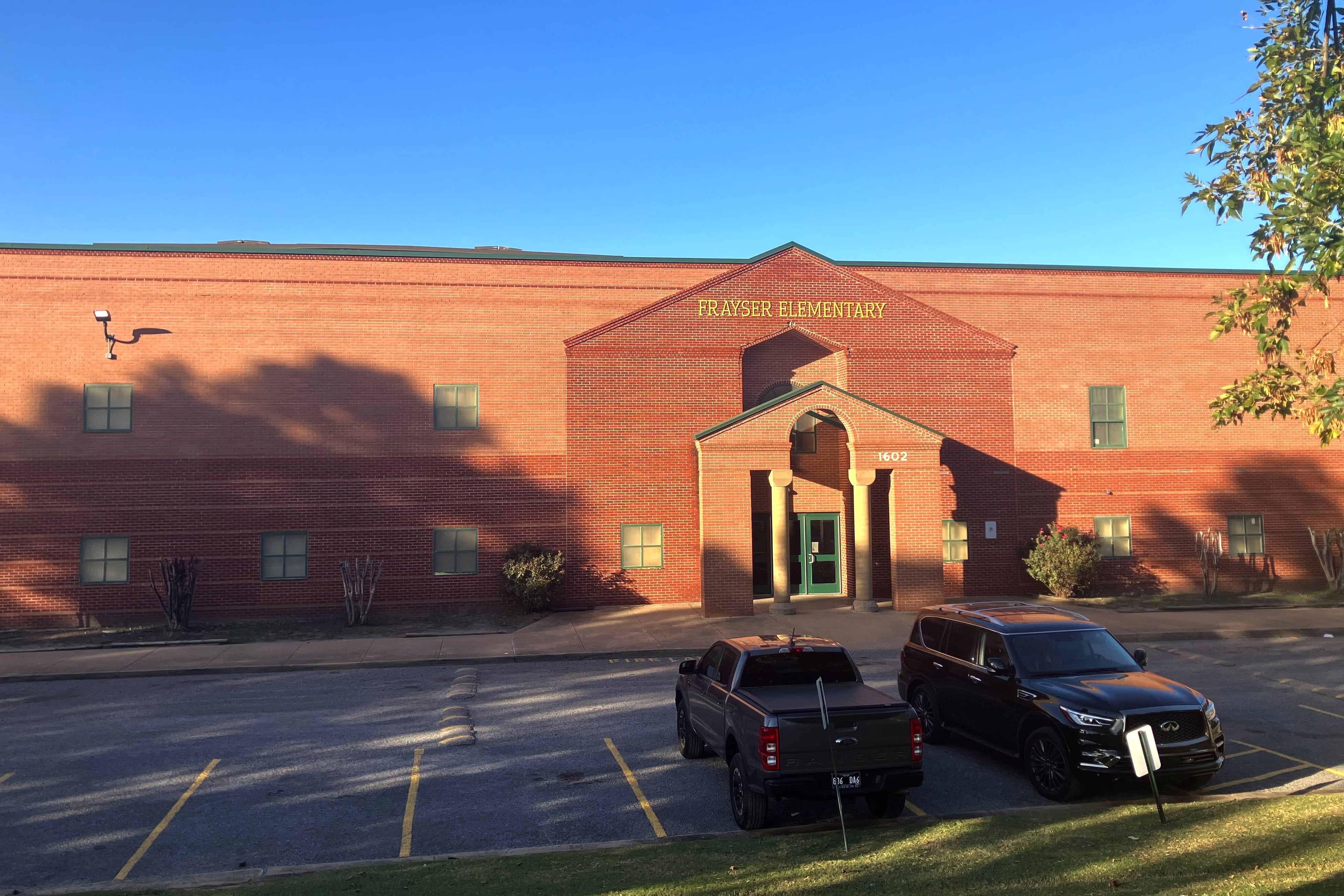 A photograph of the front of a brick school building with two cars parked in the parking lot and a clear blue sky in the background.