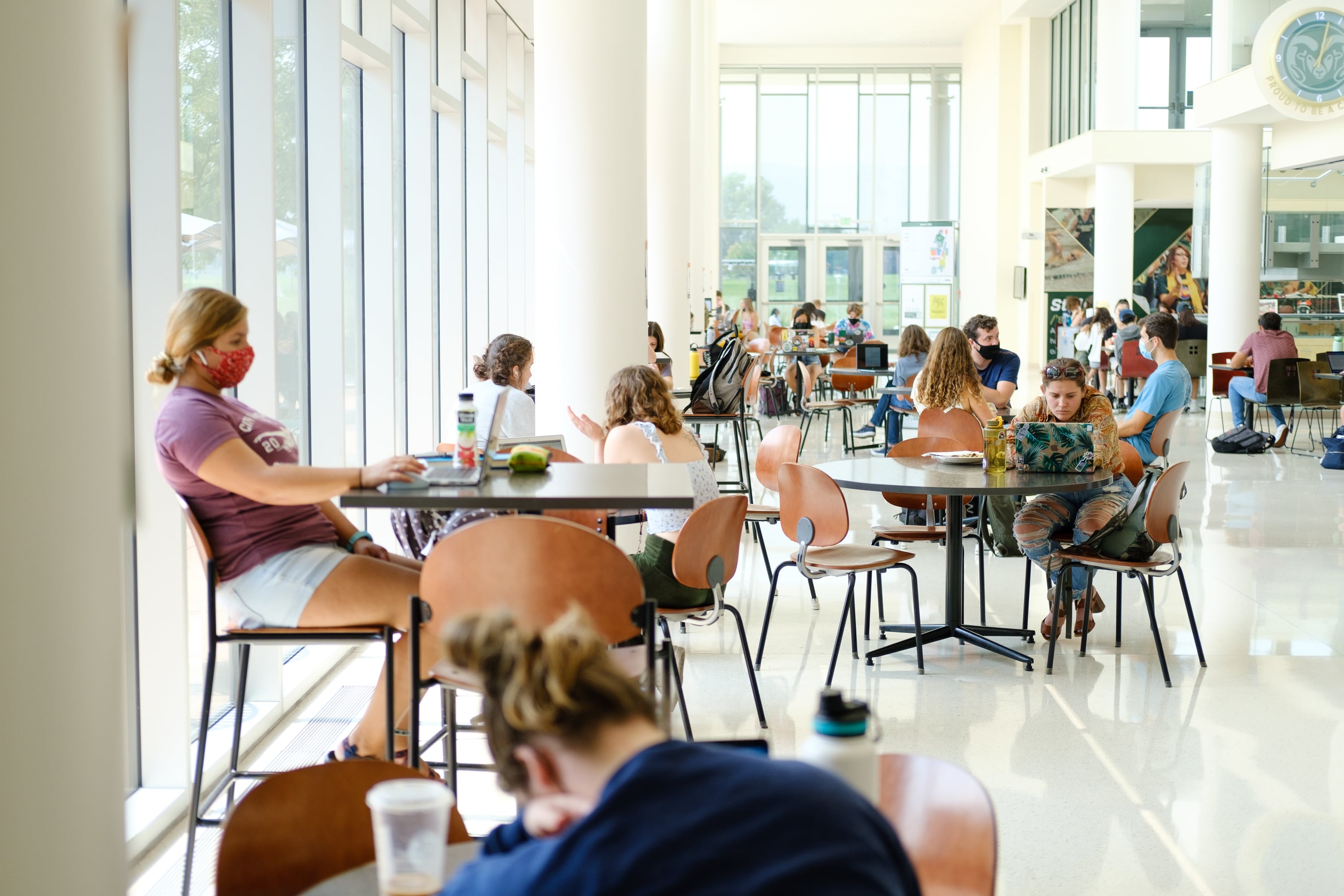 College students sit at round tables in a large room with windows.