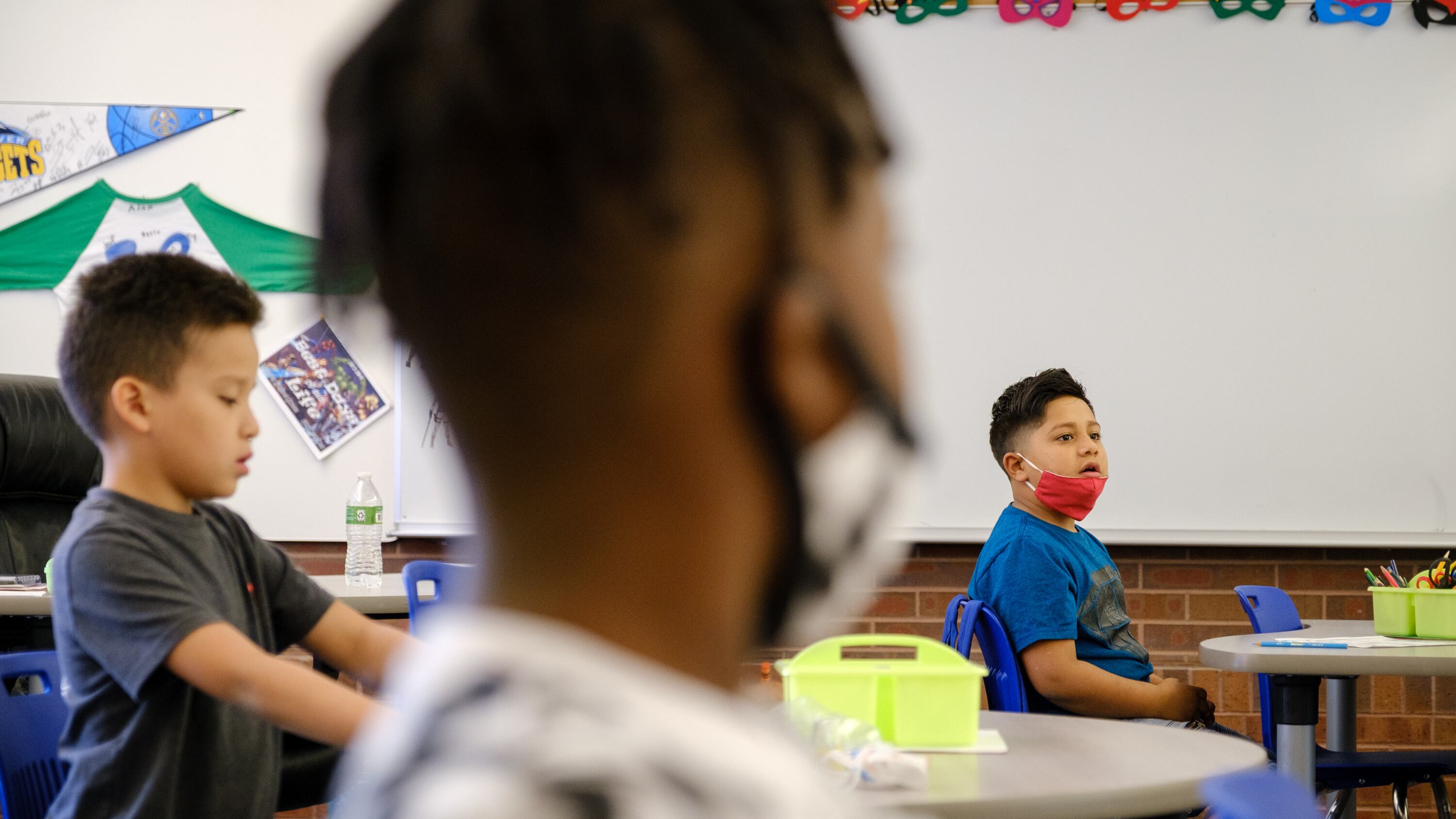 Close-up of the side of the face of a masked elementary student, sitting at a classroom desk. In the background are two other students, one wearing a mask. 