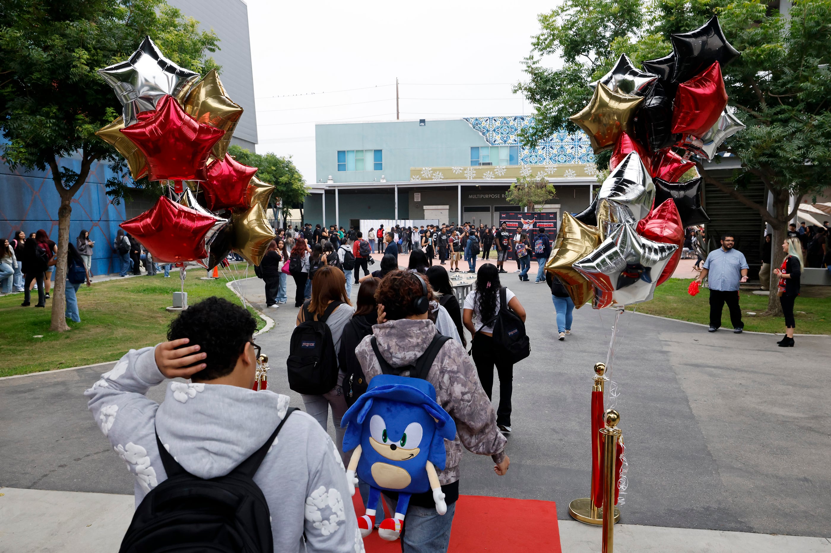 A photograph of high school students walking toward the front entrance of a school building with balloons on both sides of teh sidewalk.