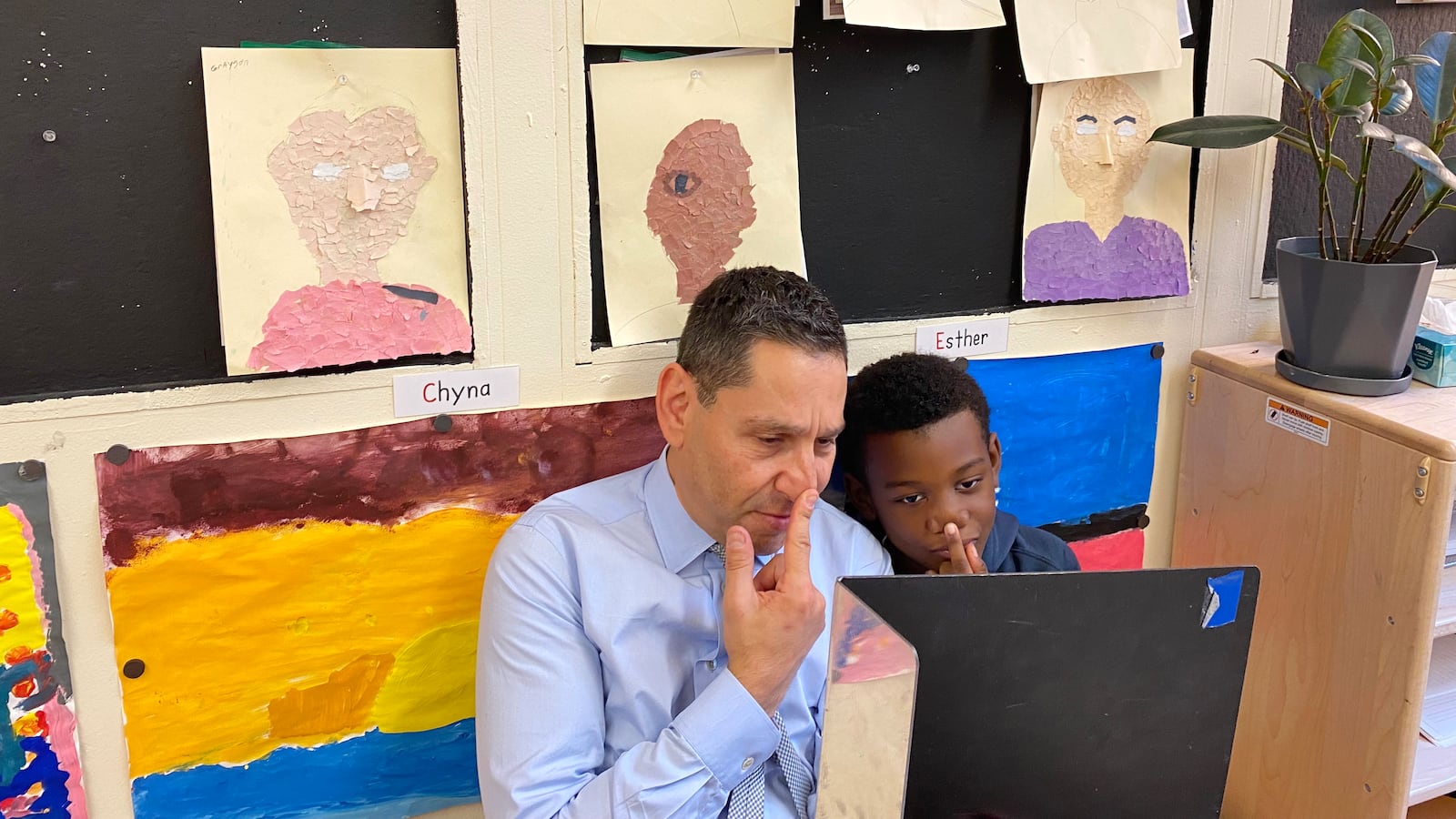 A man with a blue shirt and a student with a blue top put their fingers on their noses.