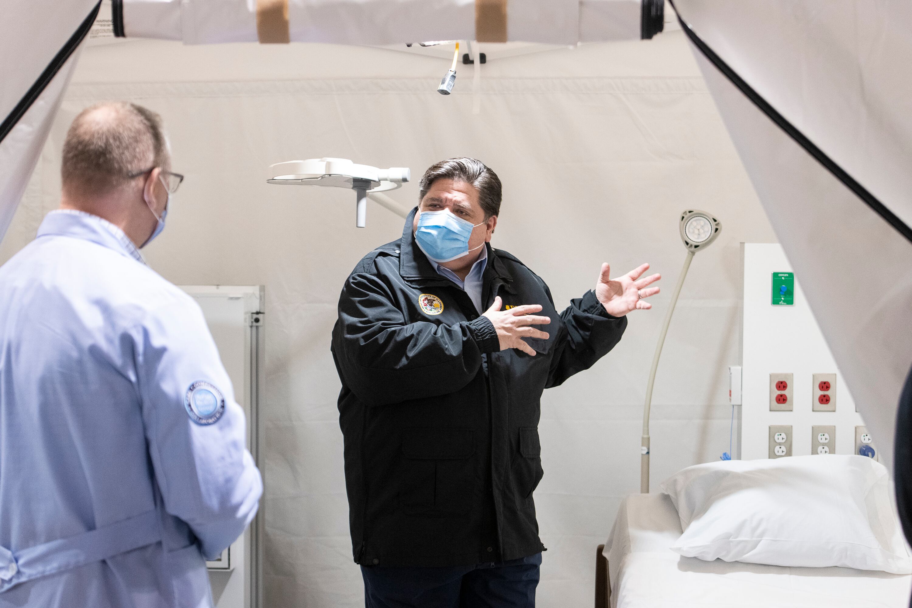 Governor J.B. Pritzker, wearing a windbreaker and a blue mask, speaks with health care worker in a white COVID treatment room.
