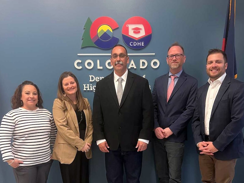 Five people in business clothes stand side by side and smile for a photograph with a grey wall in the background that has a colorful logo and the word "Colorado" in large white letters.