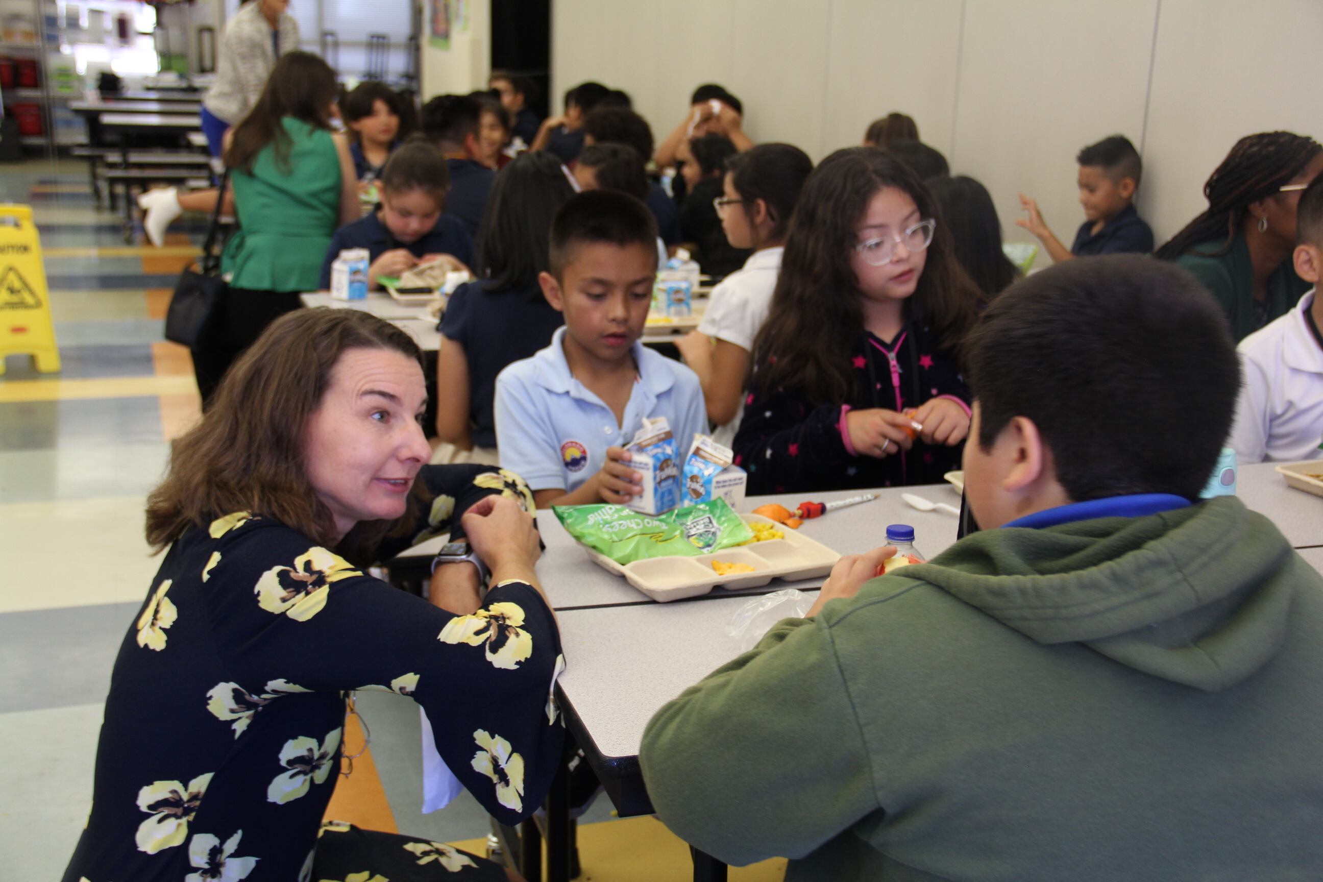 A woman with shoulder-length hair, wearing a floral print dress, leans against a cafeteria table talking to students while they eat lunch.