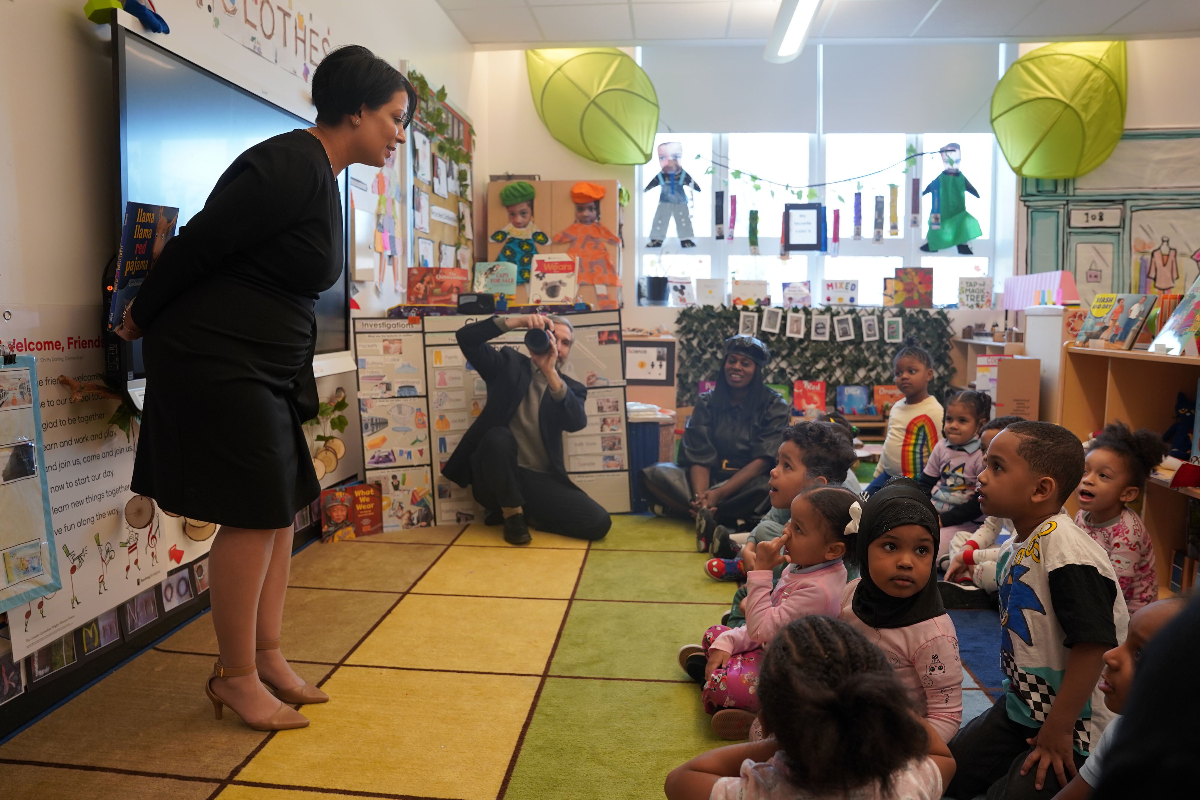 An adult woman in business clothes stands in front of a classroom full of young students sitting on a colorful rug on the floor.