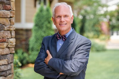 A man with white hair and wearing a bowtie and suit jacket stands with his arms crossed posing for a portrait with greenery in the background.