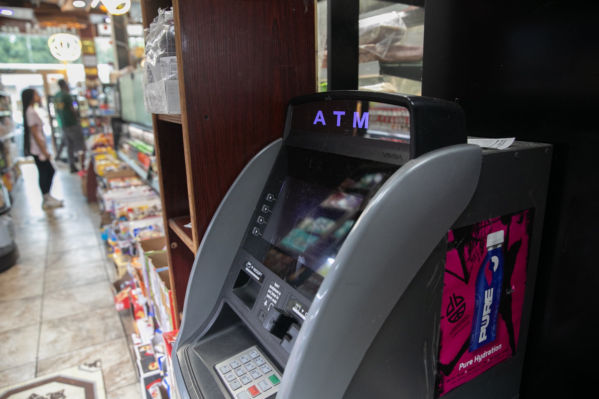 A photograph of an ATM machine inside of a bodega with two people standing in the far background.