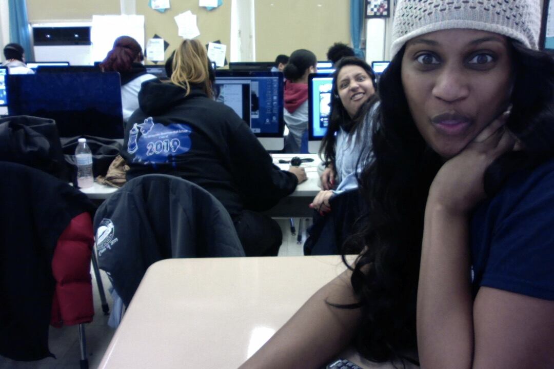 A woman in the foreground in a black T-shirt smiles at the camera while a student in the background makes a silly face.