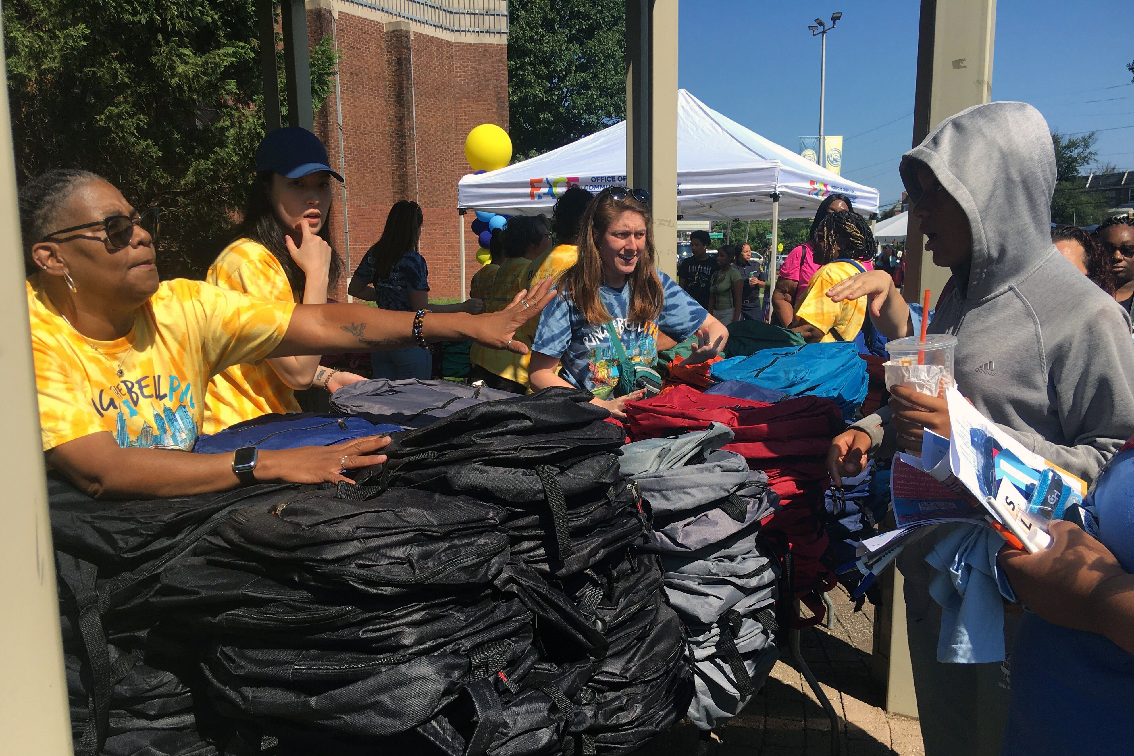 A photograph of adults standing behind a table full of backpacks outside.