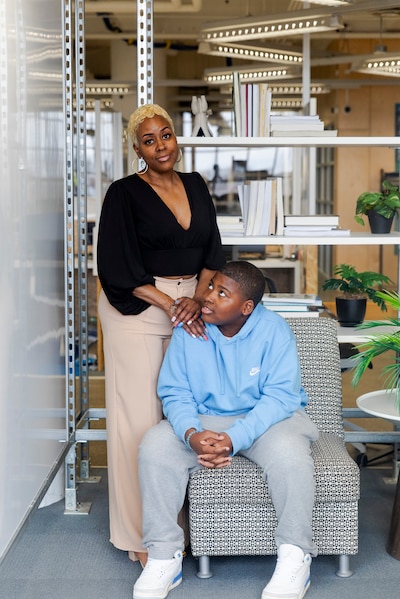 A Black mother and son pose for a portrait. The son is sitting in a chair while the mom rests her hands on his shoulder.