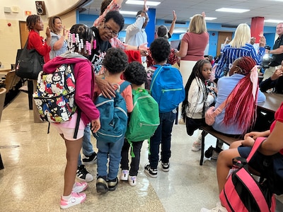 A photograph of a large group of elementary students wearing backpacks and some wearing red shirts while adults cheer them on.