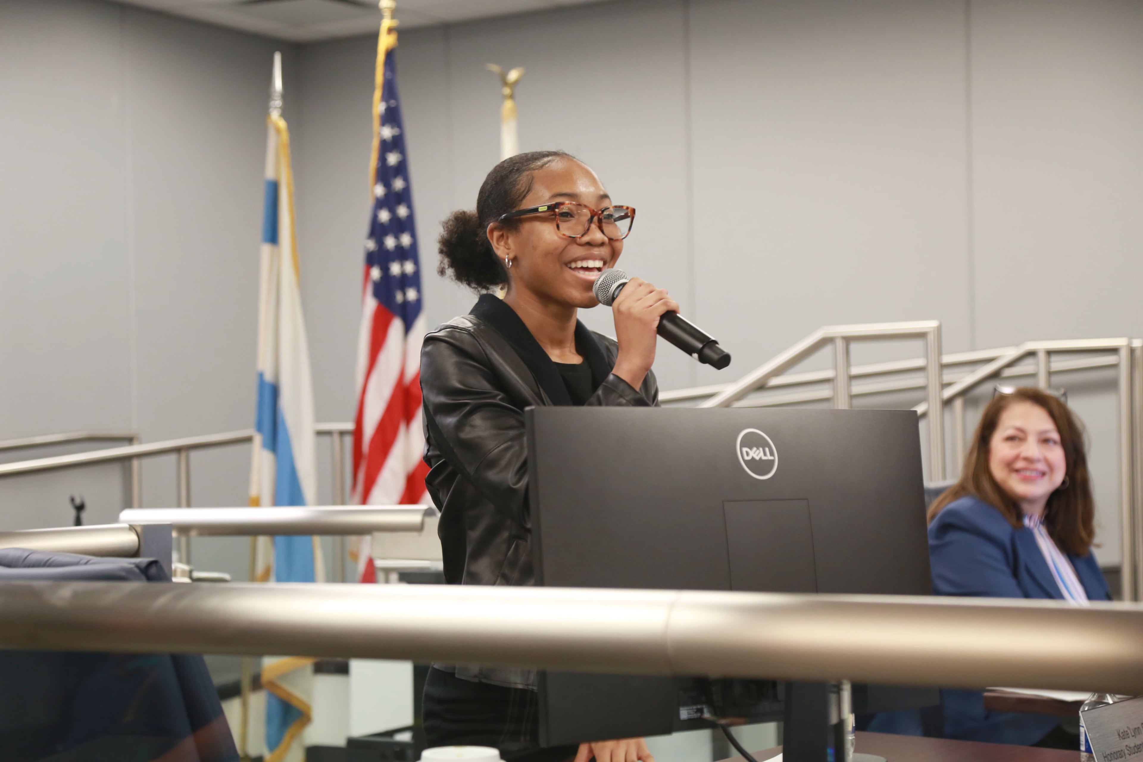 Kate’lynn Shaw, a 16-year-old girl, raises a microphone to her mouth and smiles at a school board meeting. Behind her are the Illinois, Chicago and American flags, stood up on poles. A woman to her right, sitting down, looks at her and smiles.