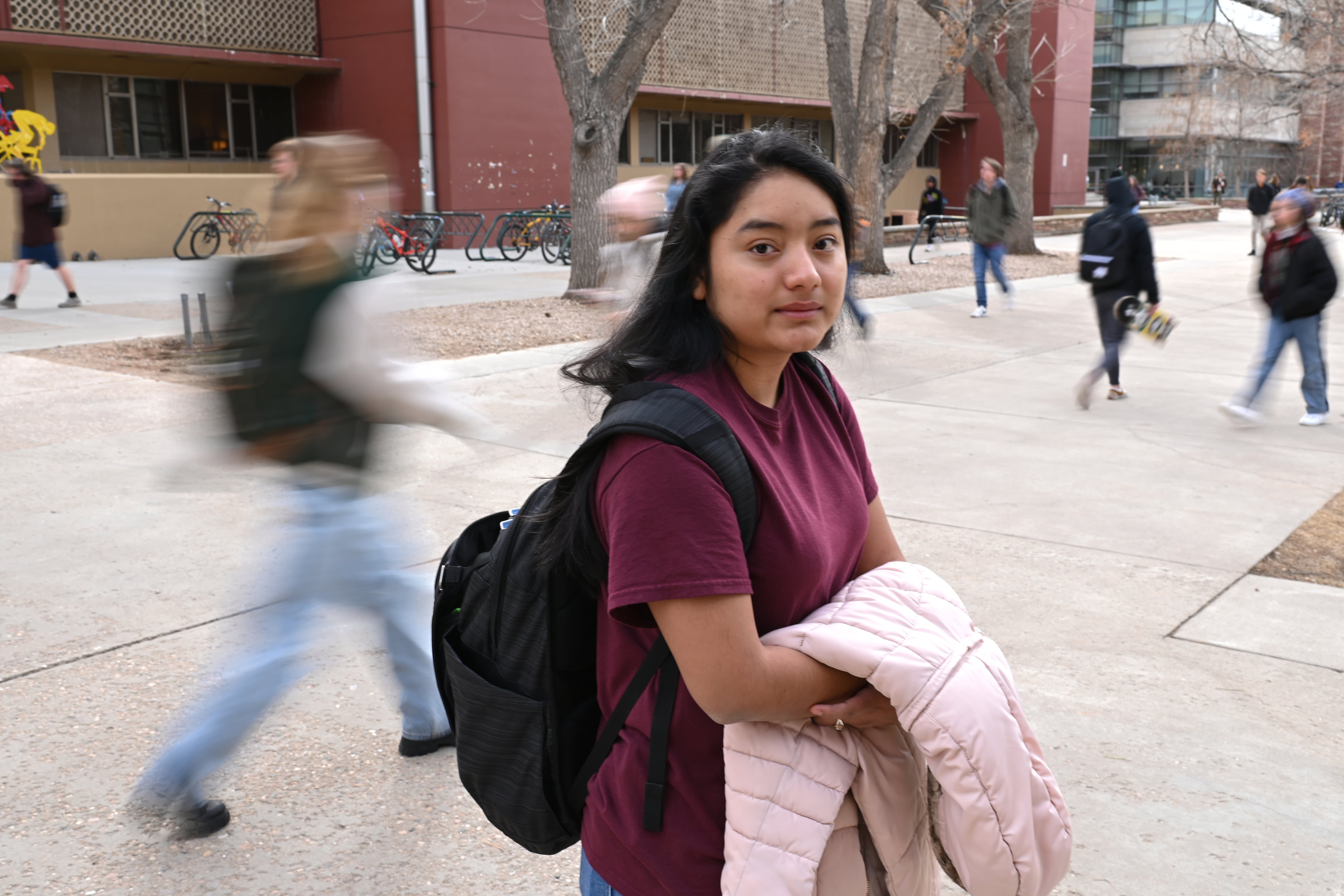 A young woman with dark hair holds a pink jacket and looks at the camera. She is standing in front of a college building and there are other blurred people walking through campus behind her.
