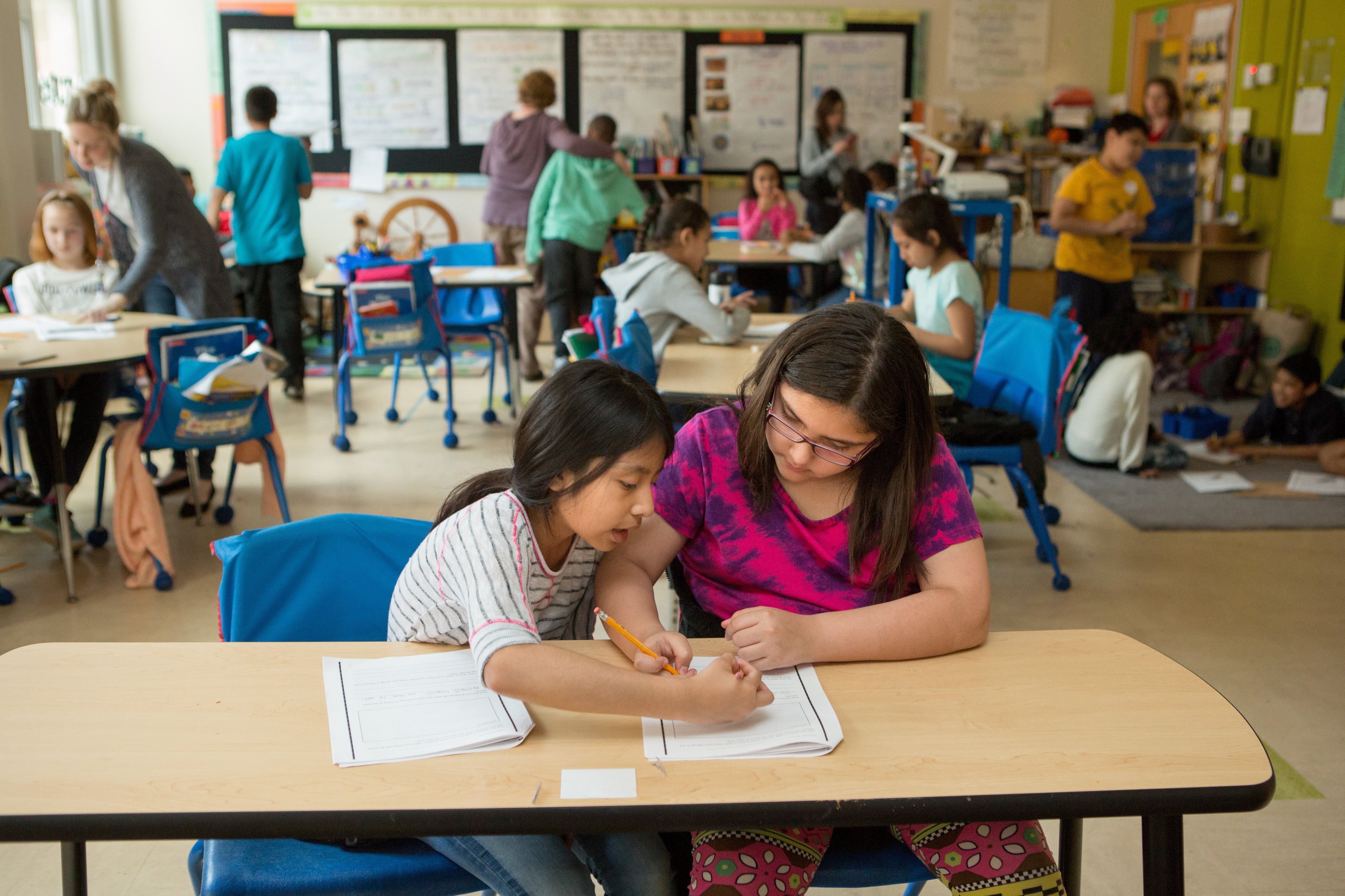 A student and an adult work at a desk while other students work in the background.