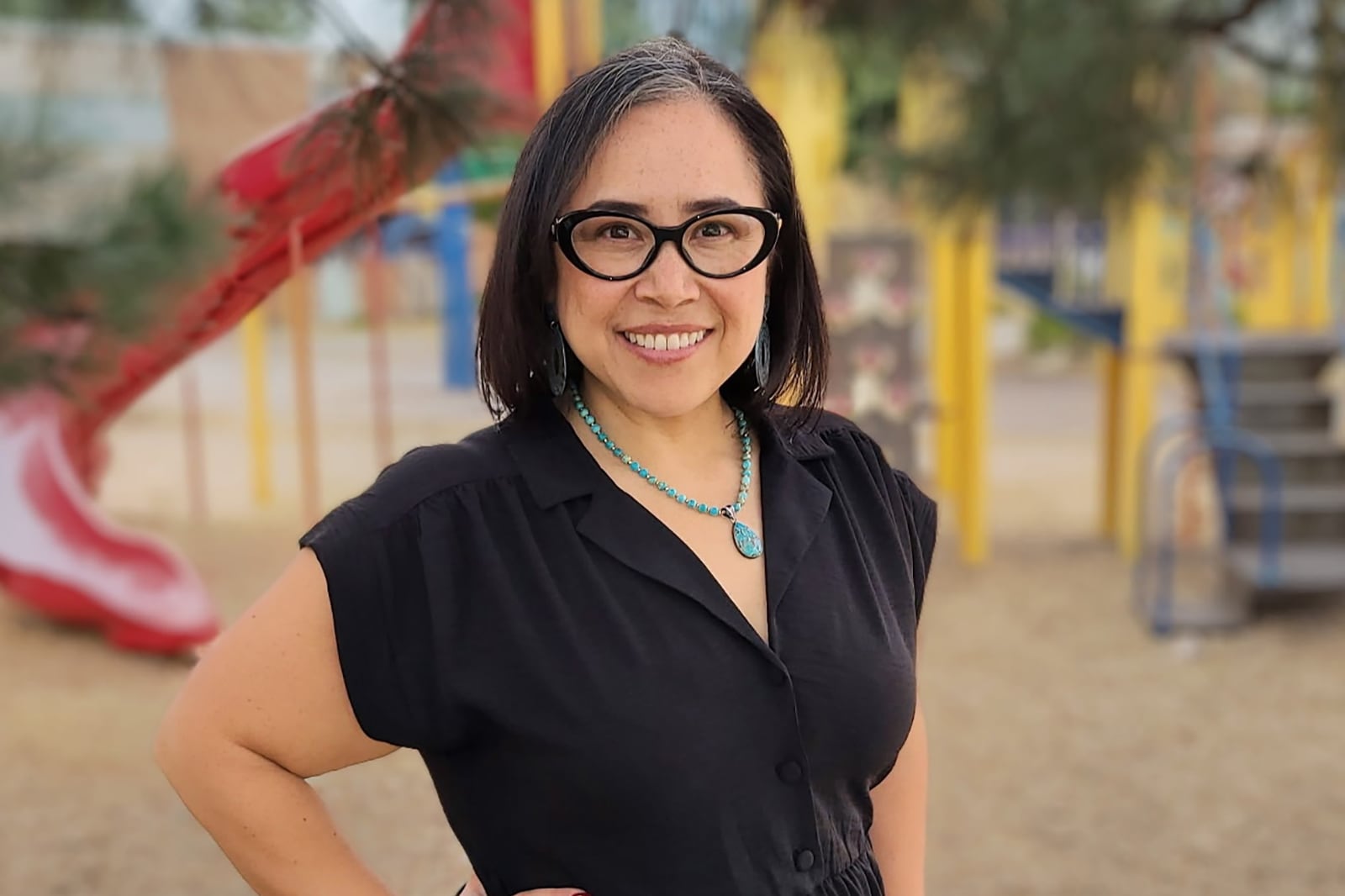 A photograph of a woman with short dark hair and wearing a black dress poses for a portrait in front of a playground.