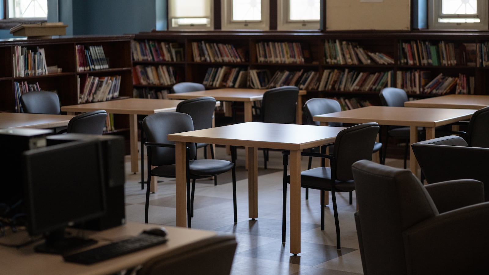 Student desks sit in an empty room at Senn High School, with bookshelves and windows in the background.