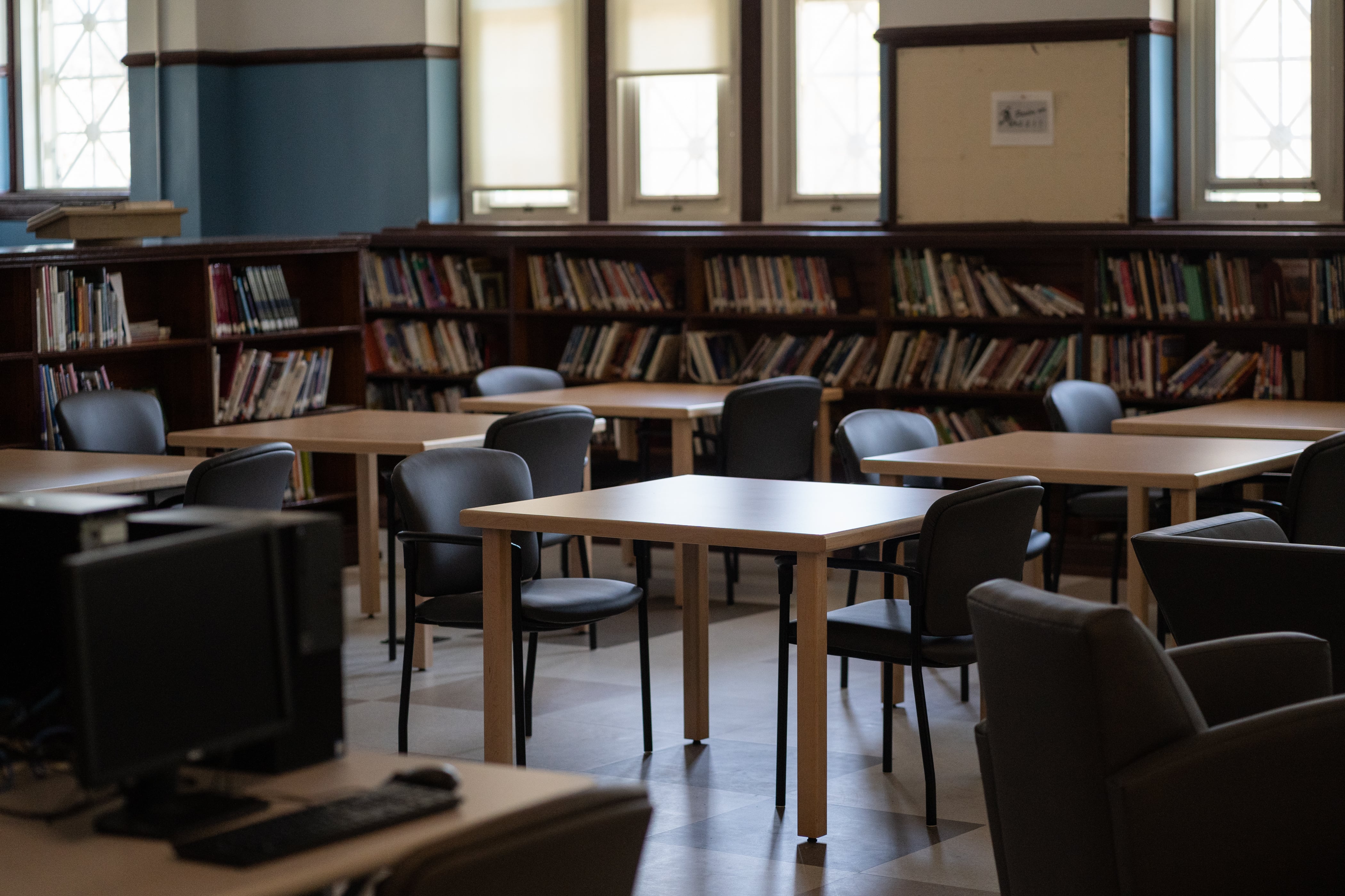 Student desks sit in an empty room at Senn High School, with bookshelves and windows in the background.