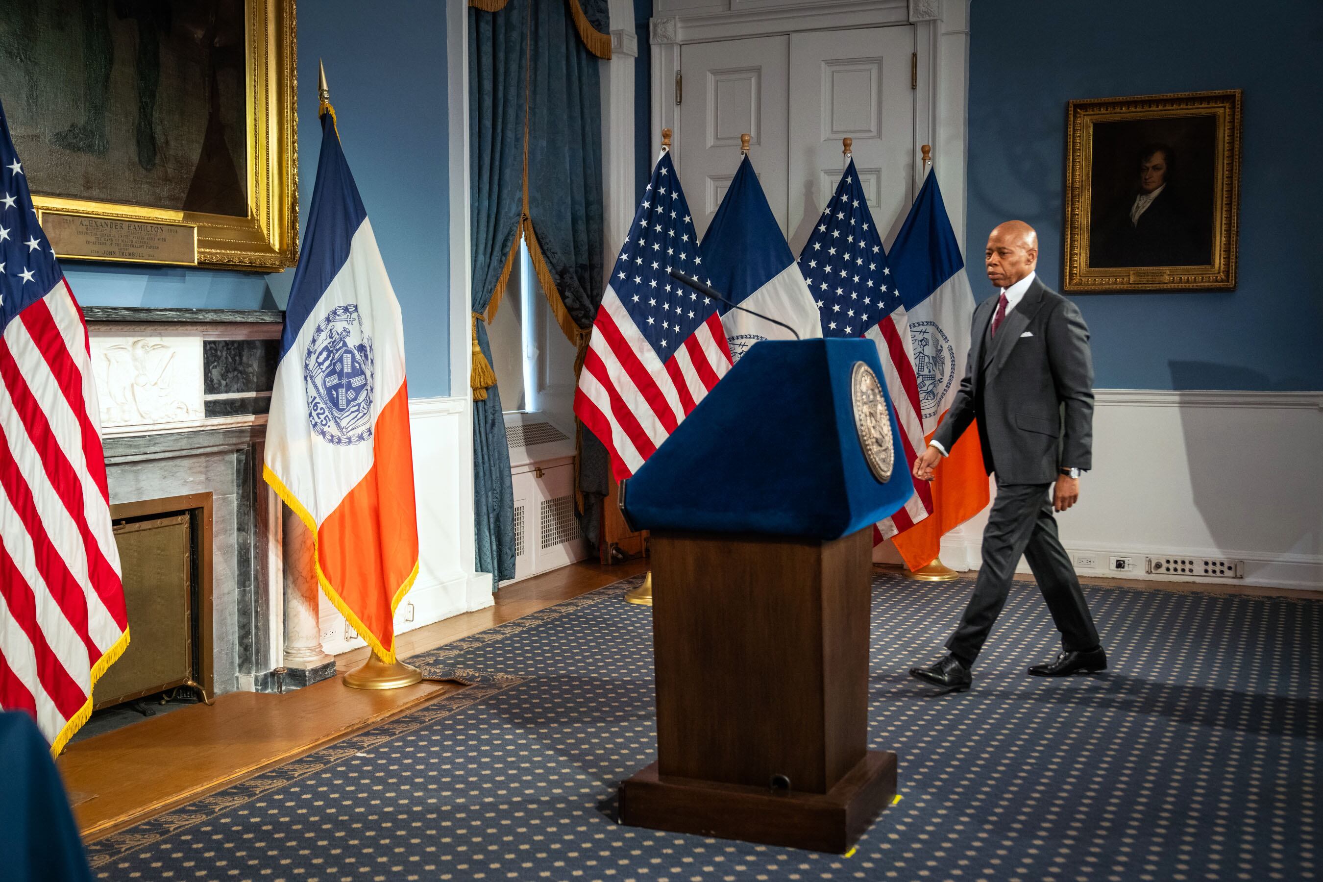 A man in a suit walks to a podium in a blue room filled with flags.