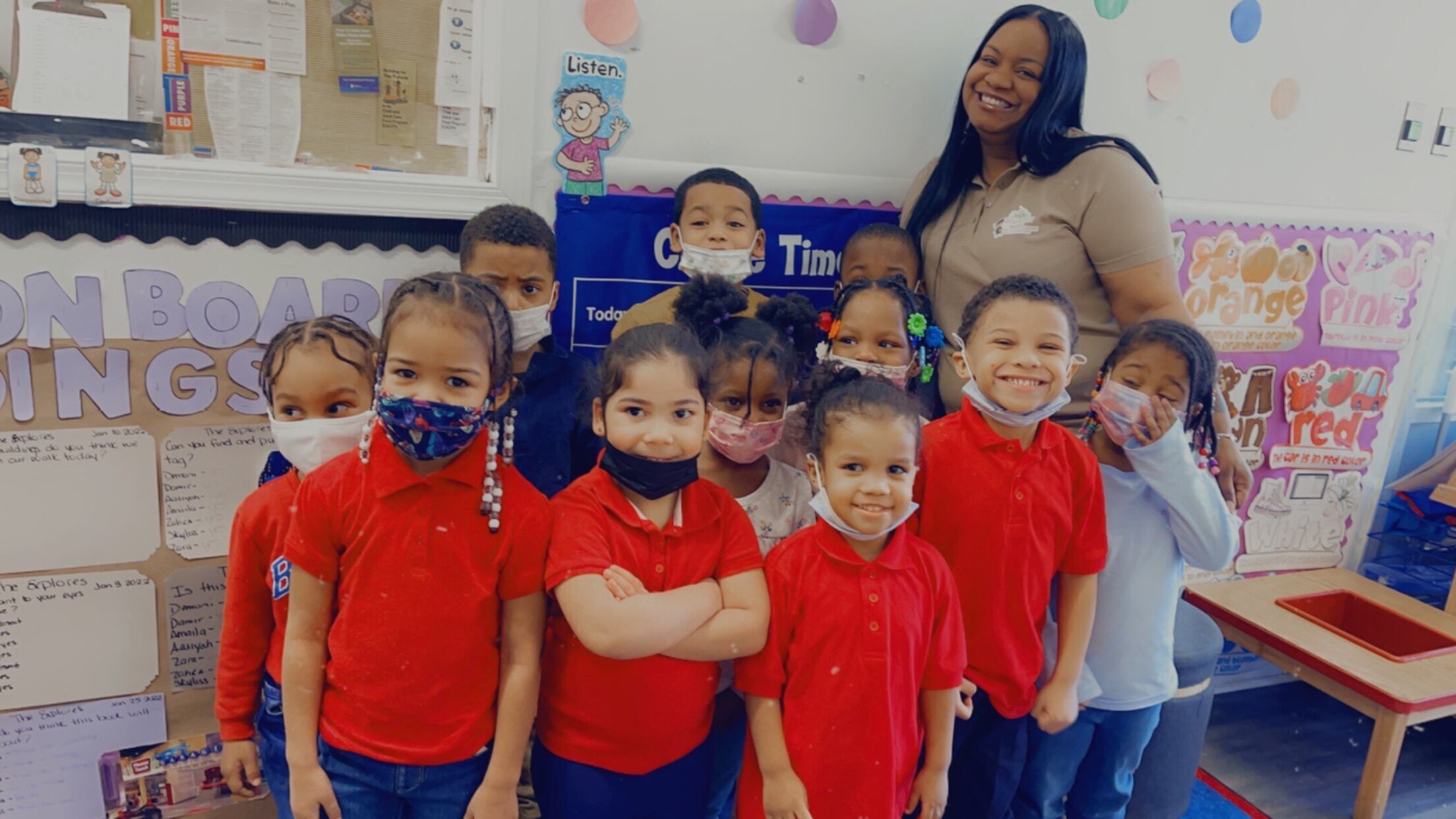 A preschool director in a brown shirt stands behind a group of children wearing red shirts.