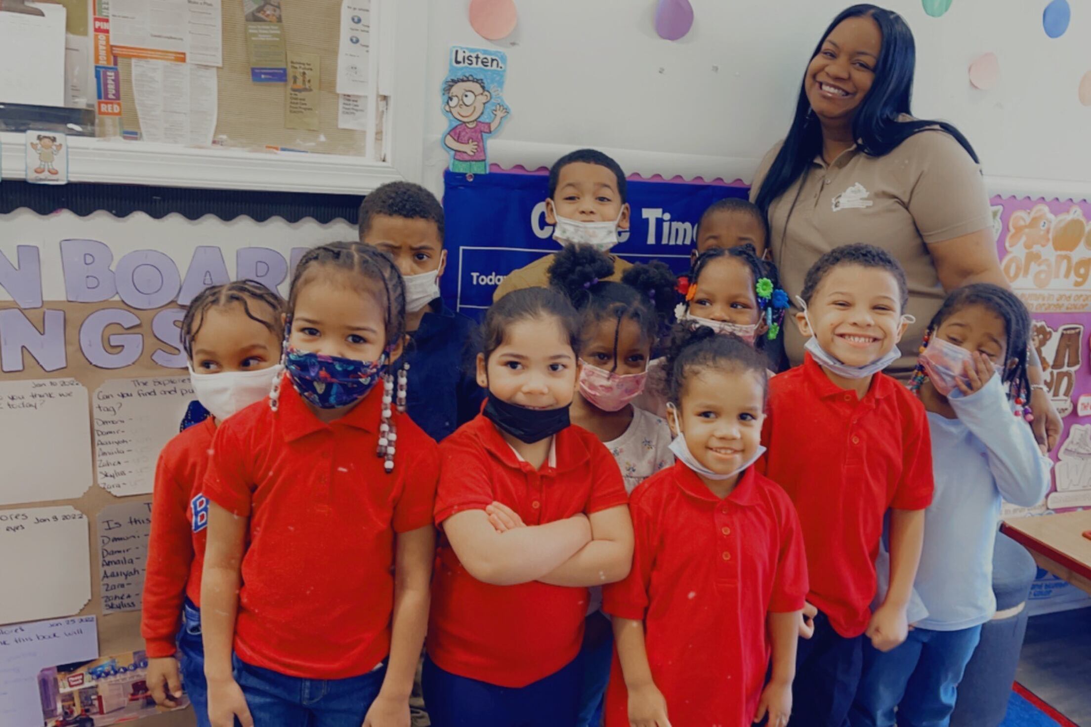 A preschool director in a brown shirt stands behind a group of children wearing red shirts.