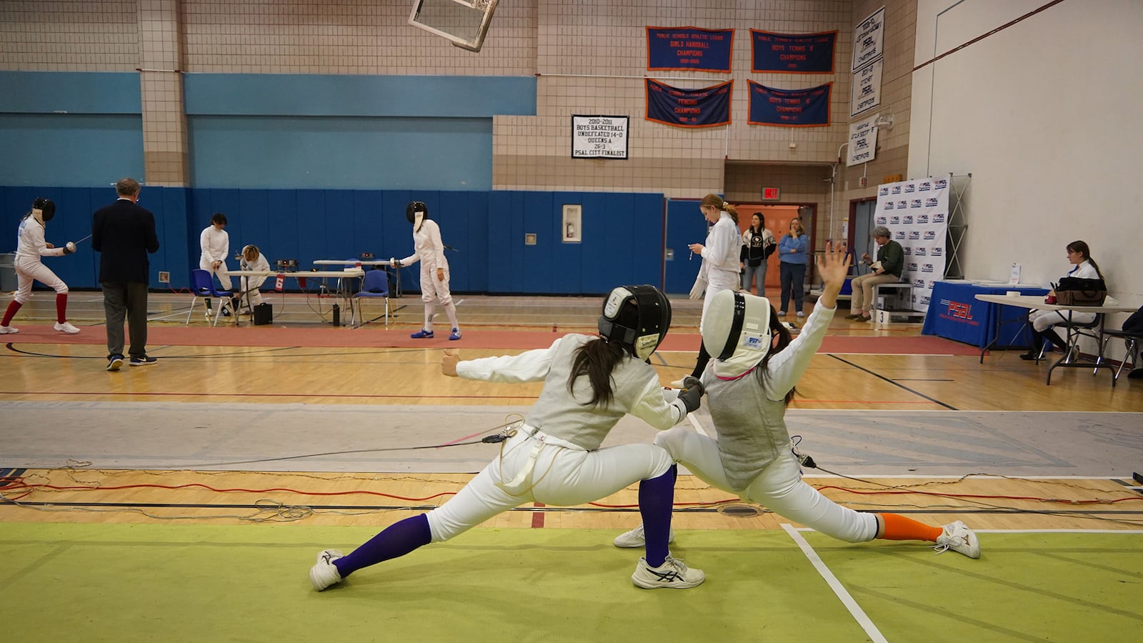 Two high school girls fencing athletes compete on a green mat in a gym with other people in the background.
