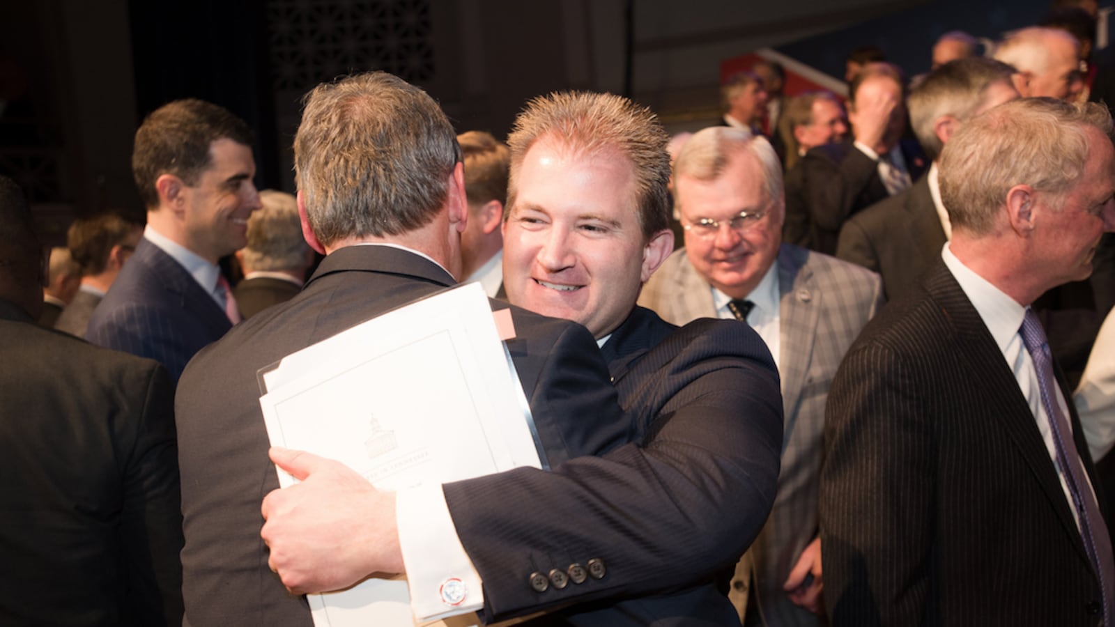 Two men wearing suits hug in the middle of a crowd.