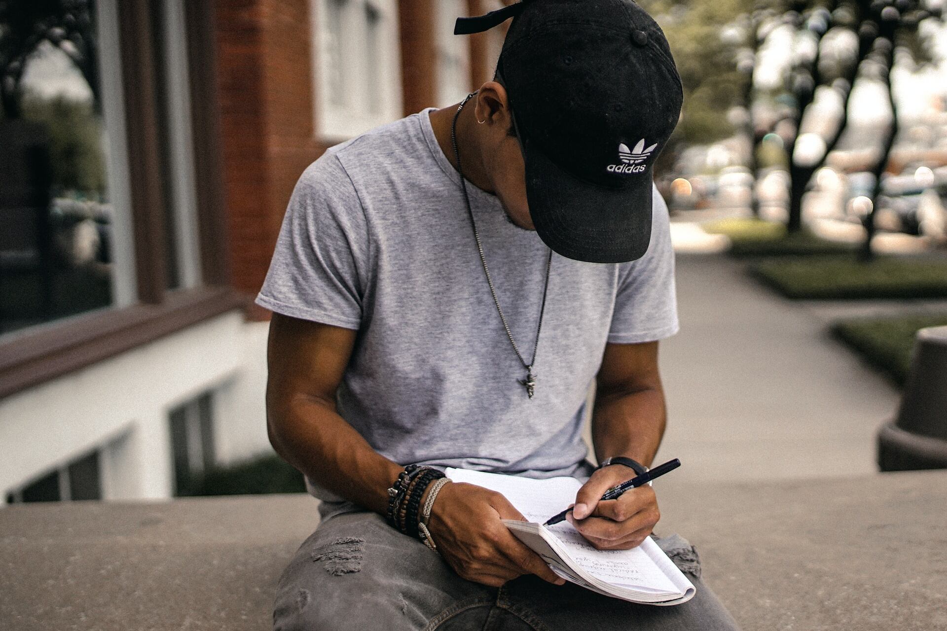 Student wearing a baseball cap sitting outside writing in a notebook.