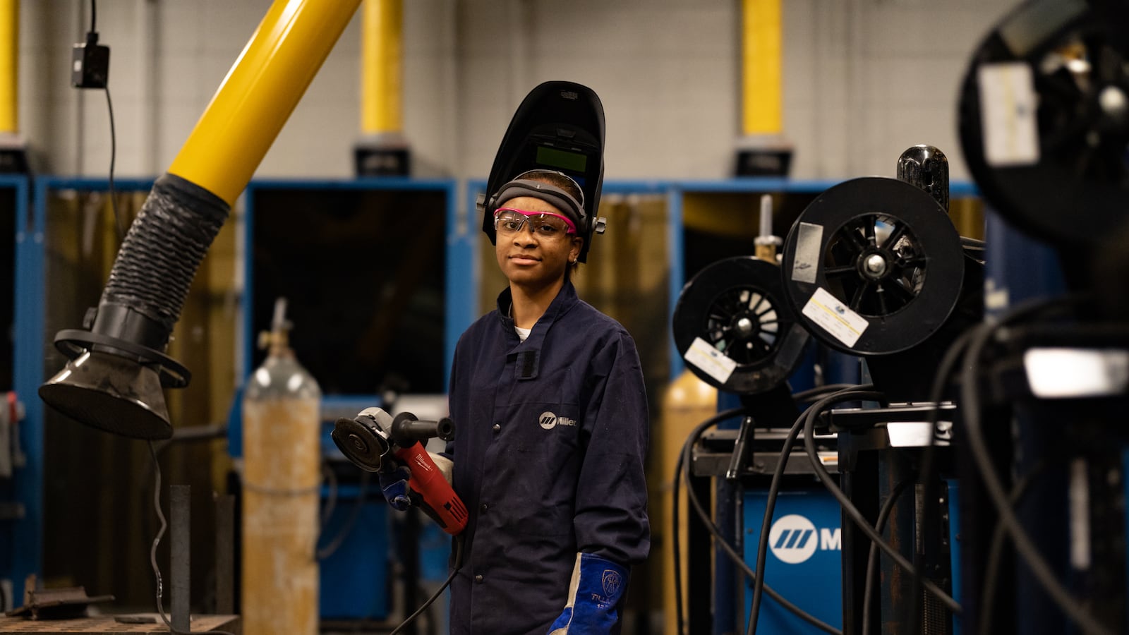 A young woman, wearing protective gear for welding, stands in a technical career classroom and poses for a portrait.