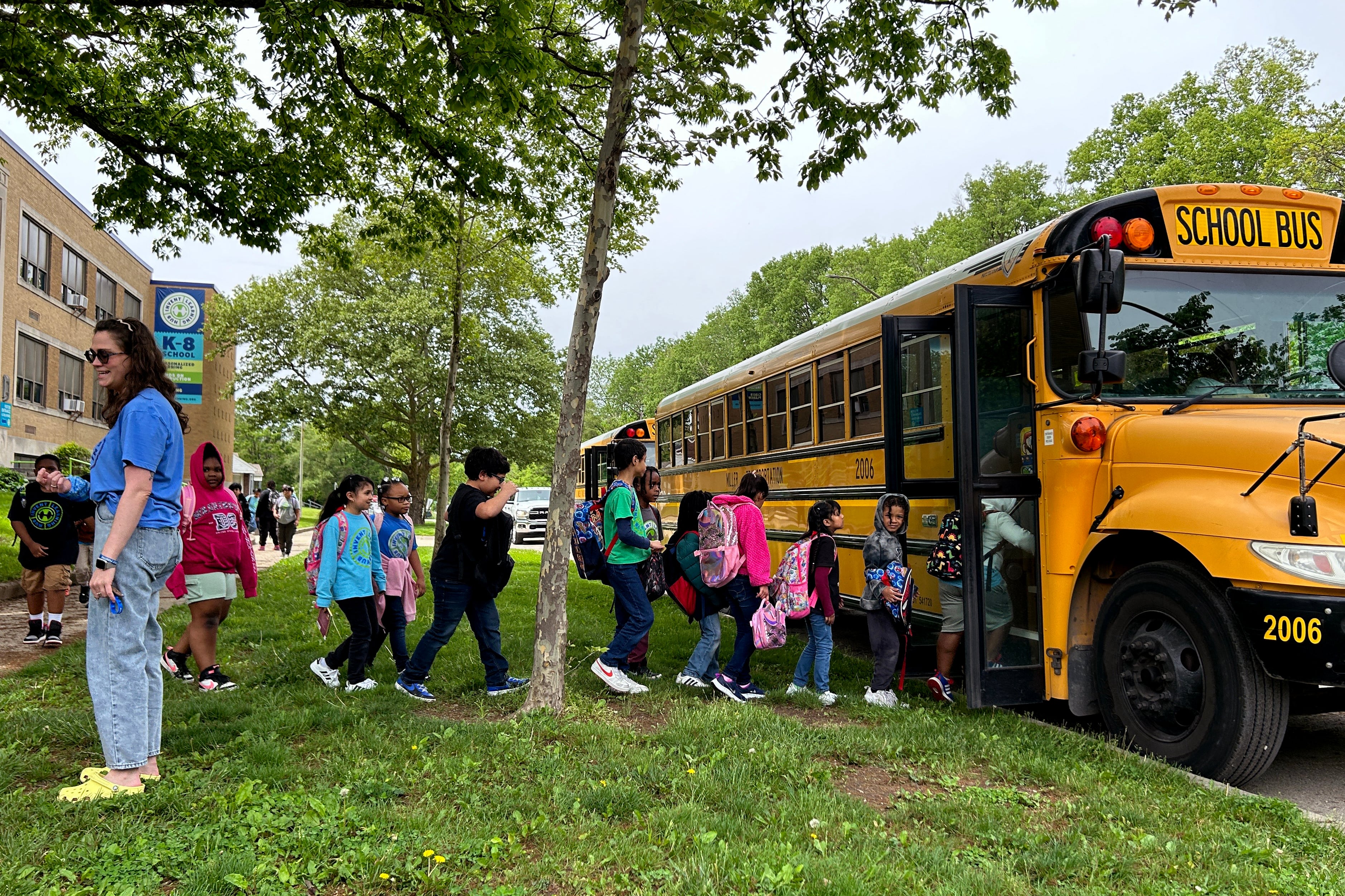 A group of young students line up outside with two yellow and black school busses near by.