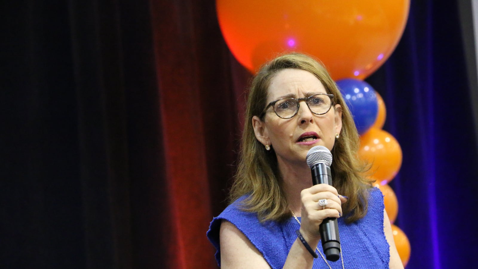 A woman with medium length brown hair, glasses and wearing a blue blouse, holds a microphone while speaking in front of balloons and a backdrop.