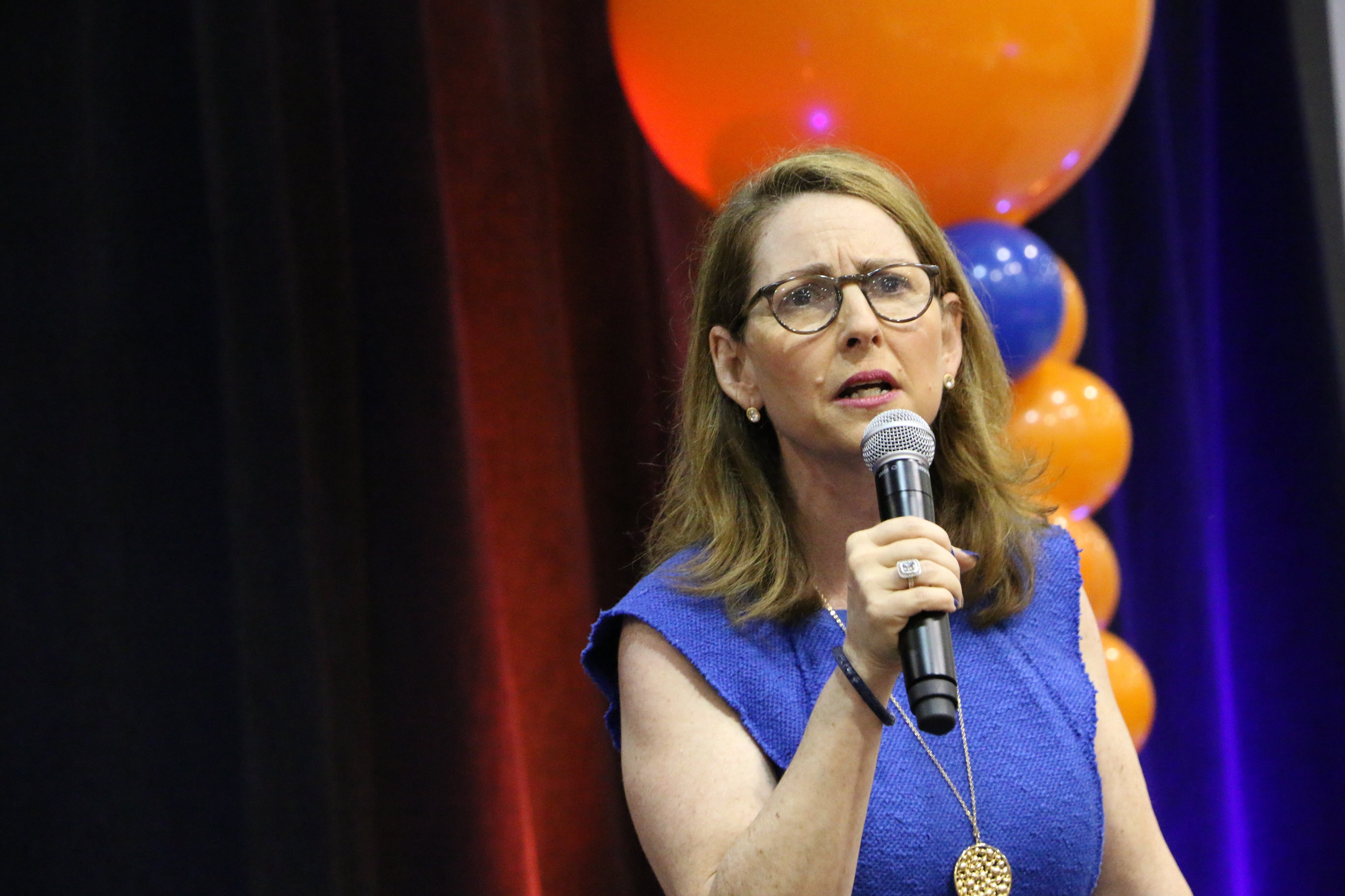 A woman with medium length brown hair, glasses and wearing a blue blouse, holds a microphone while speaking in front of balloons and a backdrop.