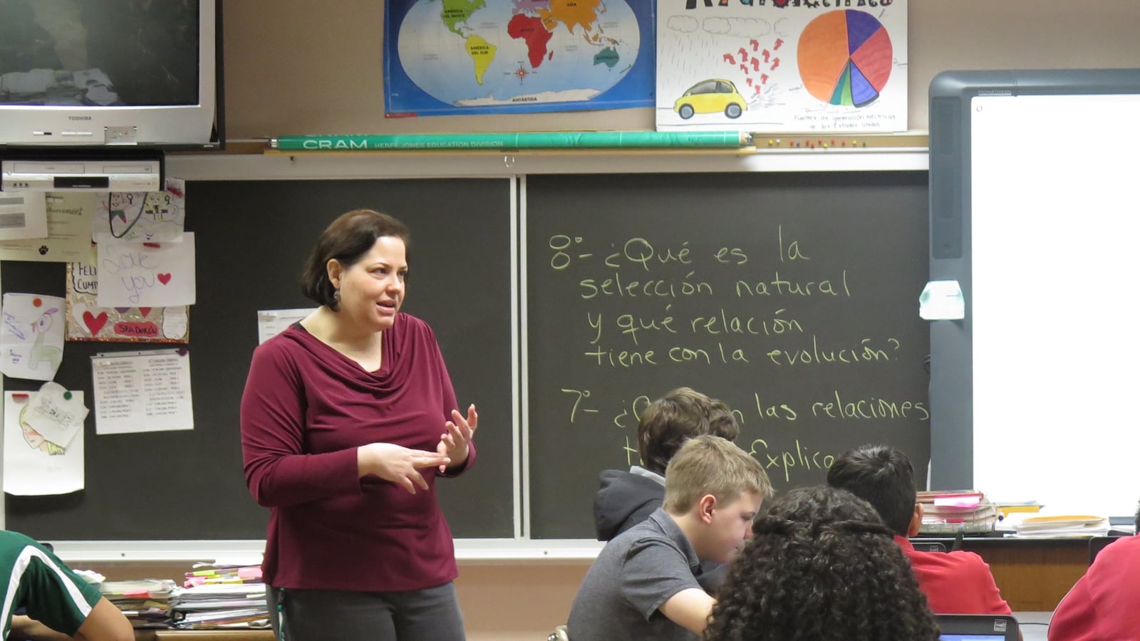 A woman in a maroon blouse instructs her class, standing in between the desks of her students