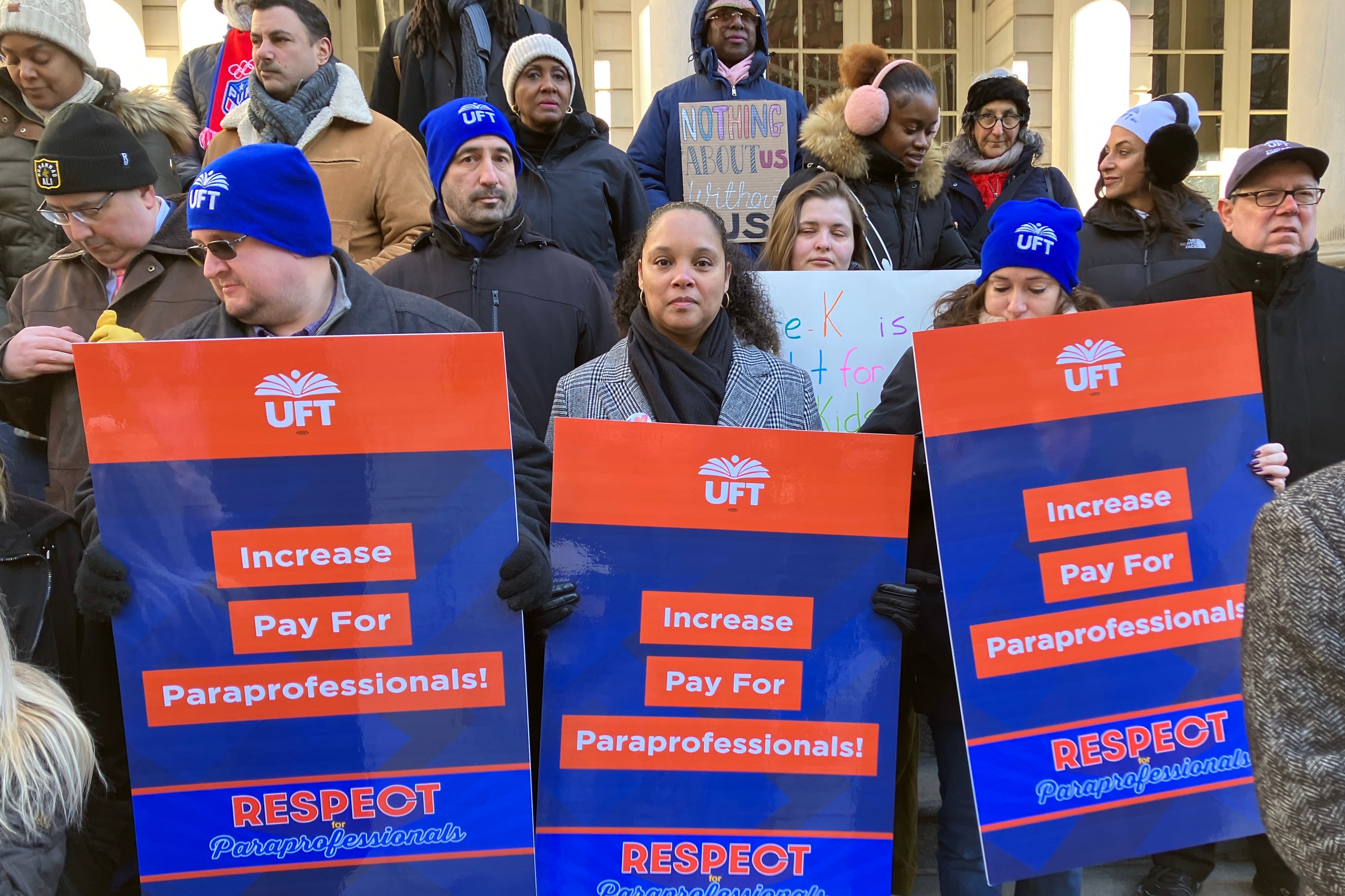A group of adults hold signs and wear jackets outside of a large stone building.