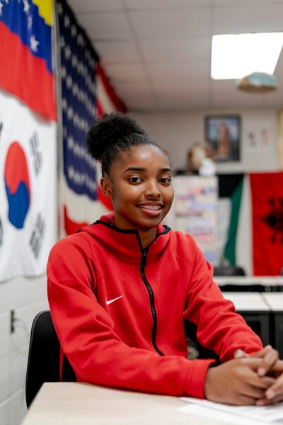 A high school student wearing a bright red sweater poses for a portrait while sitting at a desk in a classroom.