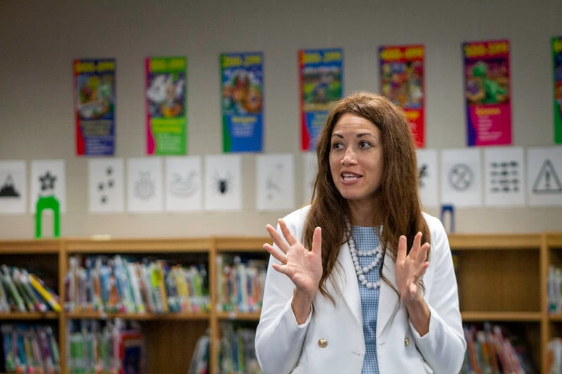 A woman stands in a school library and gestures with her hands.