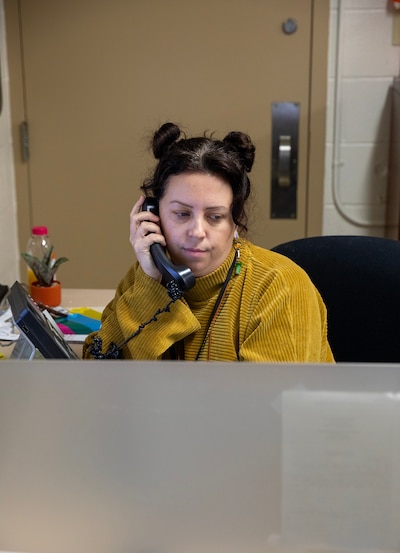A woman with brown hair in two buns and wearing a yellow sweater talks while holding a phone at a desk with a bookshelf and a door in the background.