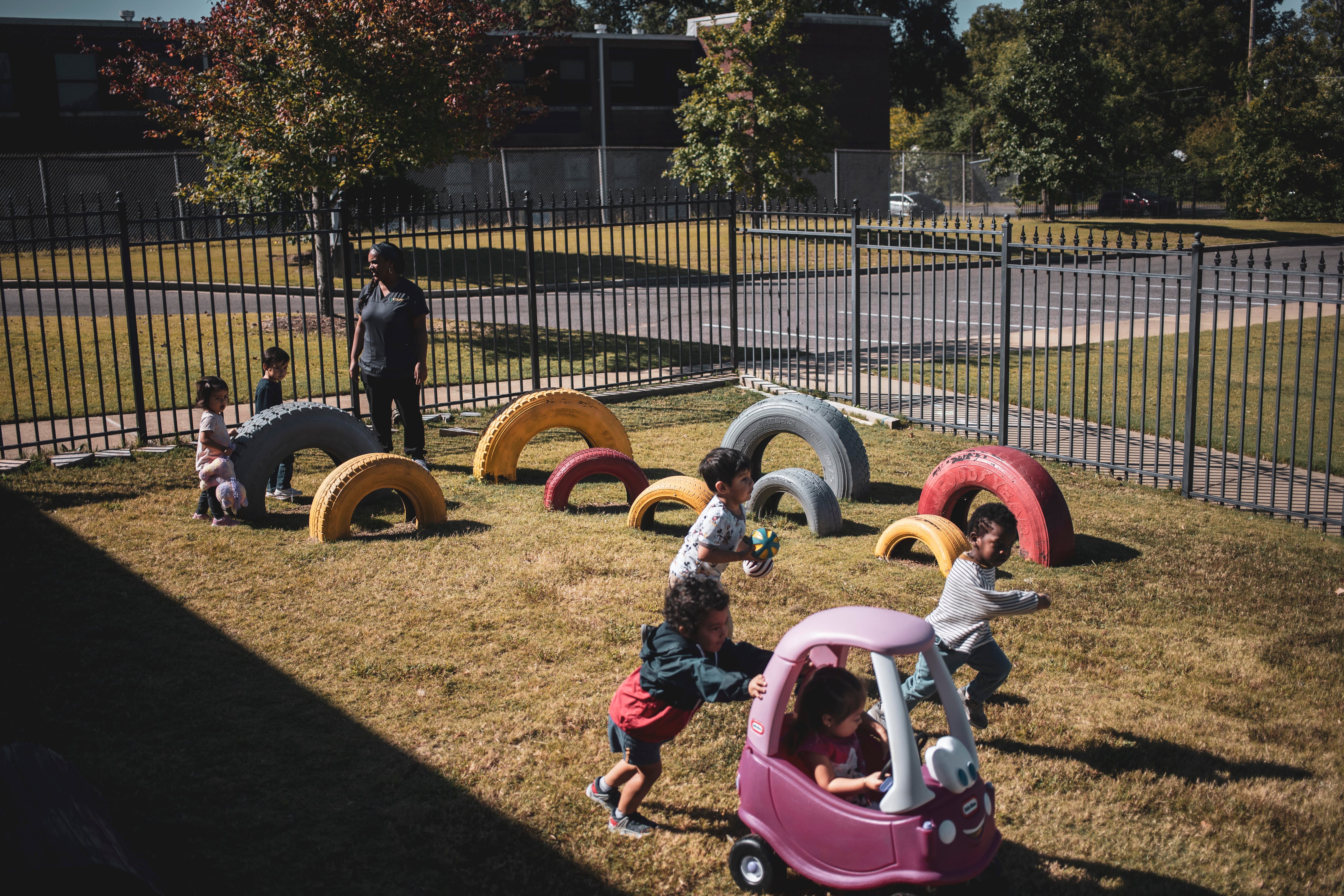 Children play in a fenced tire playground with one of their teachers. In the foreground, a boy pushes a girl in a pink car.