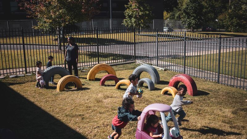 Children play in a fenced tire playground with one of their teachers. In the foreground, a boy pushes a girl in a pink car.