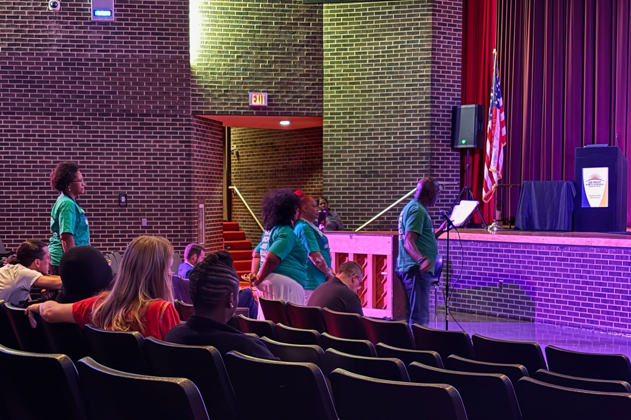 A man speaks into a microphone at a Detroit school board meeting, with several others lined up to speak and a few people sitting in the audience.