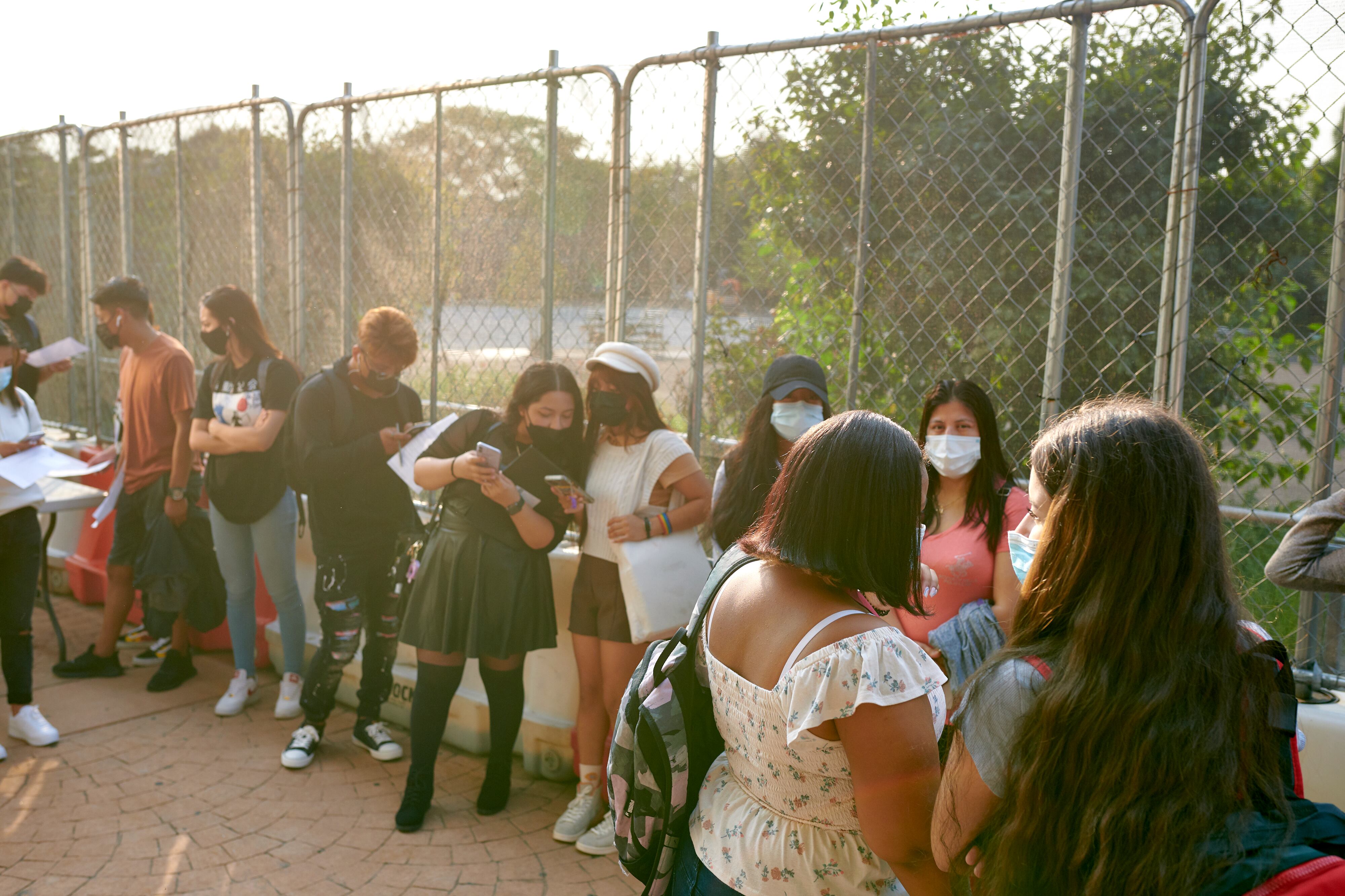 A line of students wearing masks outside of a school with a ray of sun.