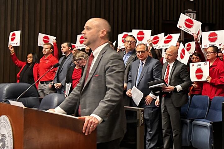 A man in a gray suit stands at a podium. People in red holding signs are behind him.